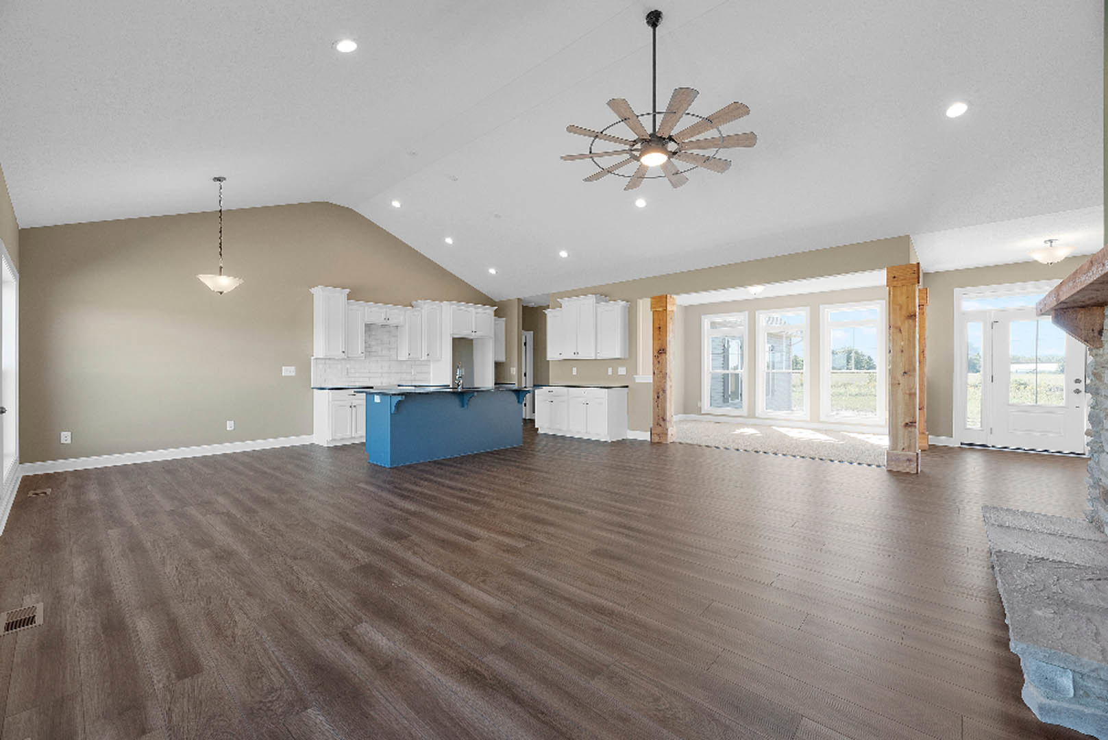 Spacious open-plan room featuring wood flooring, white walls, blue accent wall, ceiling fan with light, kitchen island with metal trim, and large windows.