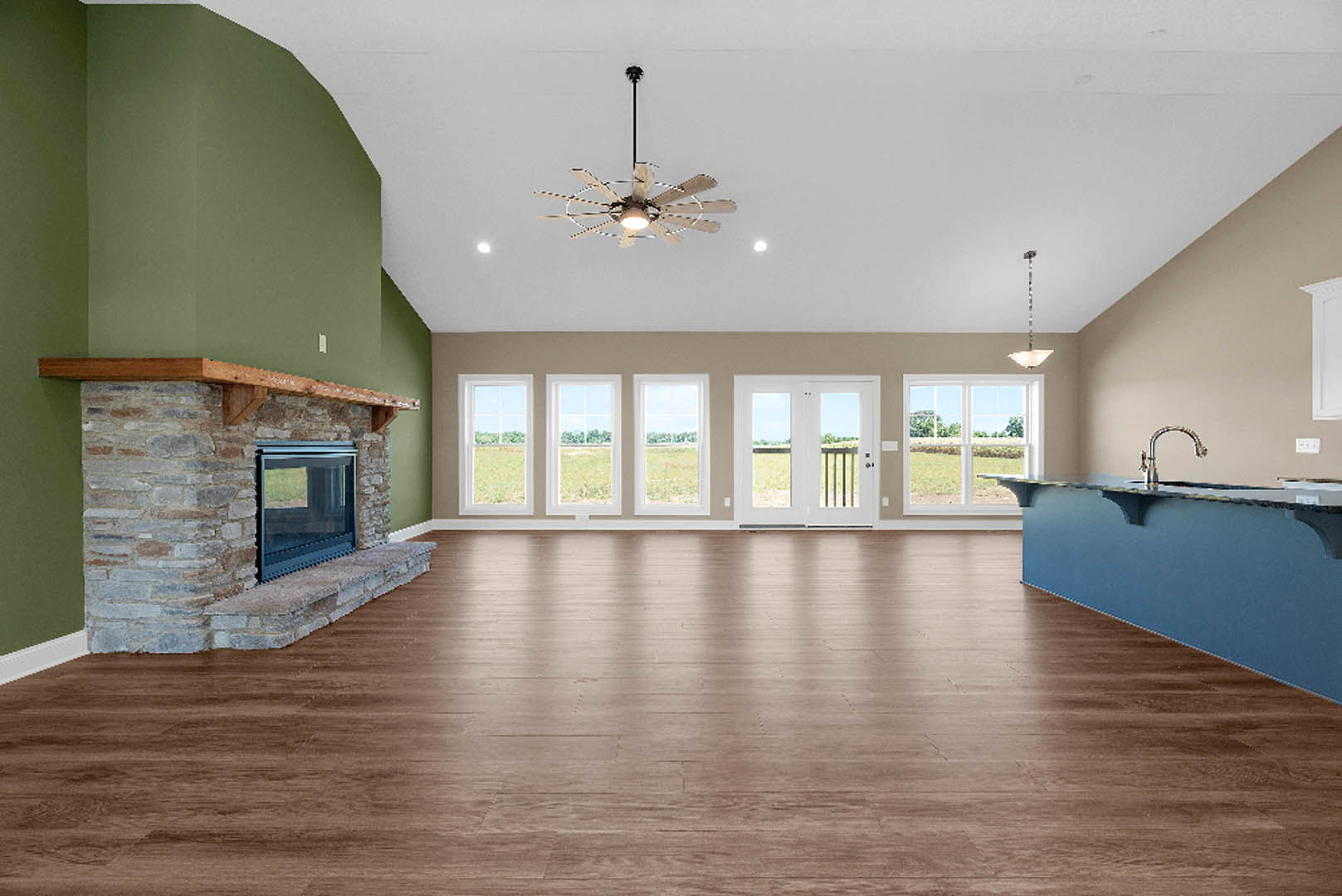 Spacious living room featuring hardwood floors, white walls, large windows, a central fireplace with stone surround, ceiling fan with light fixture, blue bar area with faucet, and