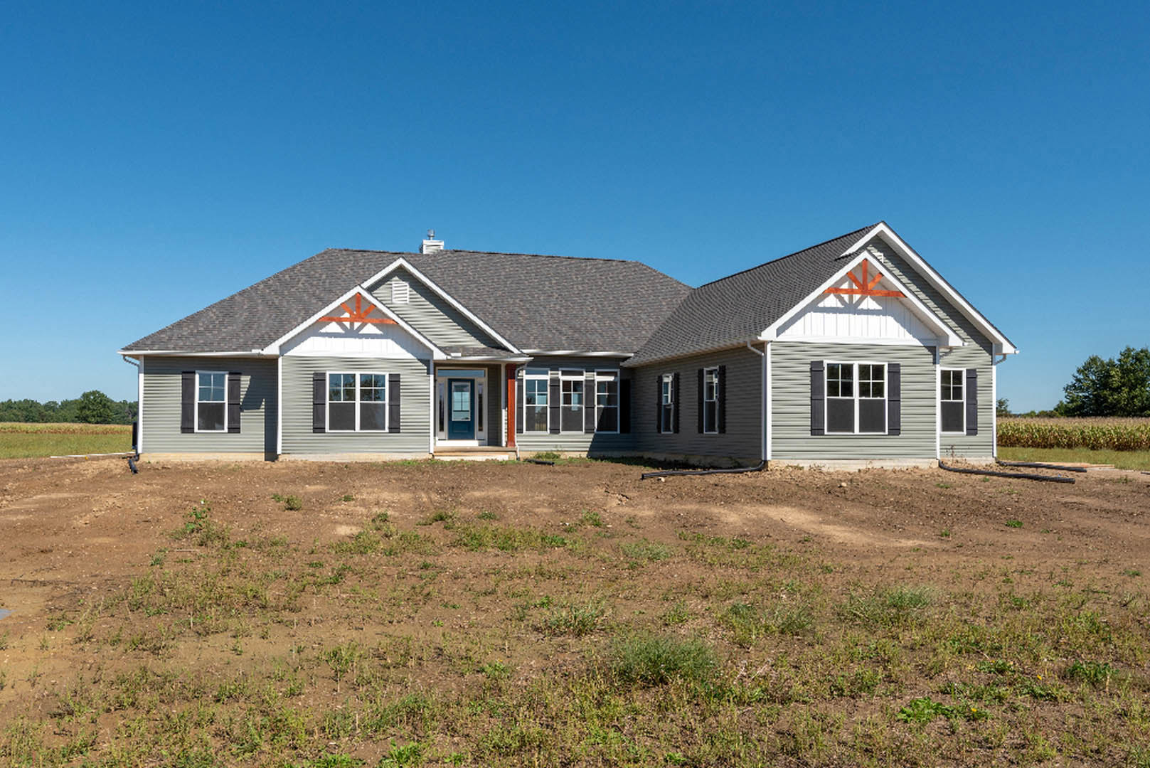 Two-story house with blue front door, white-trimmed windows, and dirt field with patches of grass under clear blue sky