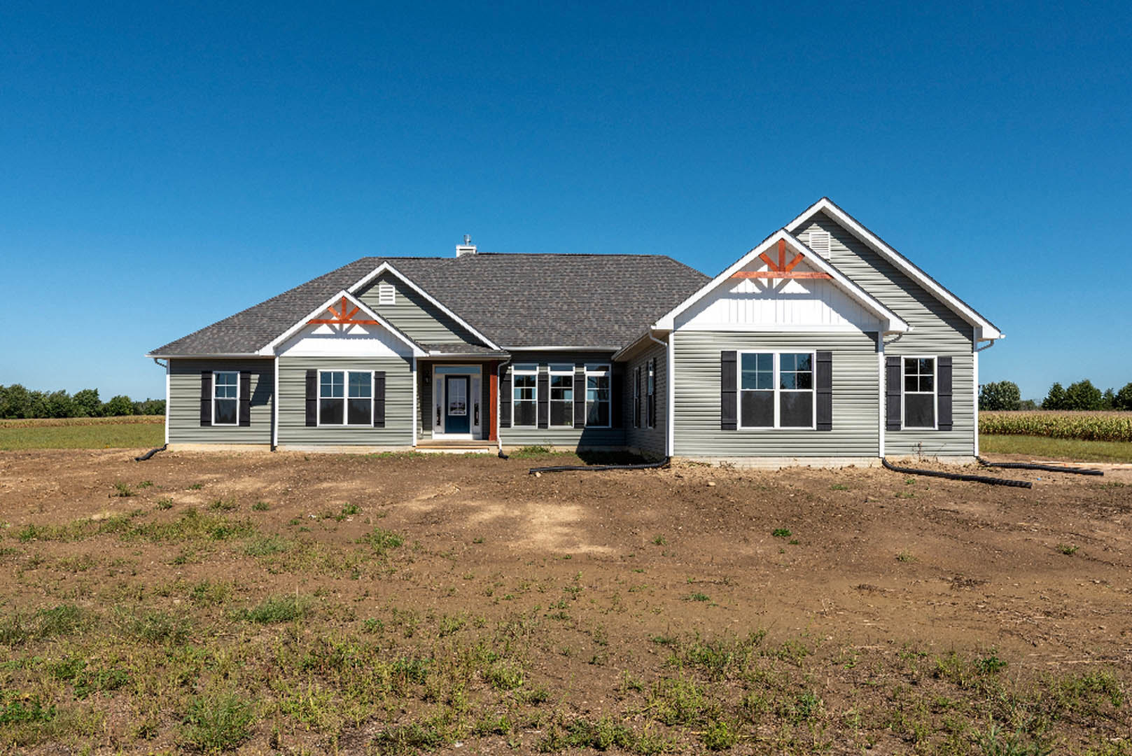 Modern single-story house with light siding and large windows, set on a dirt lot under a clear blue sky, garage visible to the side, minimal landscaping
