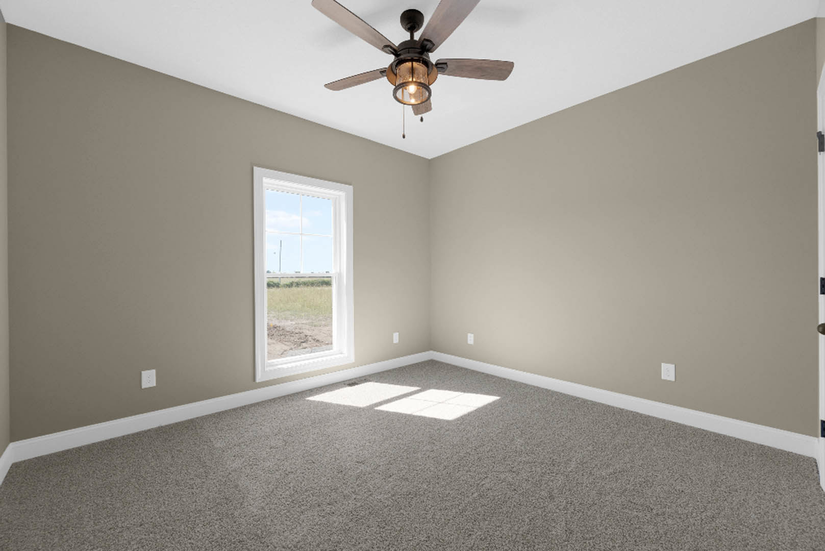 Neutral-toned room with carpet flooring, white walls, ceiling fan with light fixture, large window overlooking grassy field, sunlight casting shadows across floor.