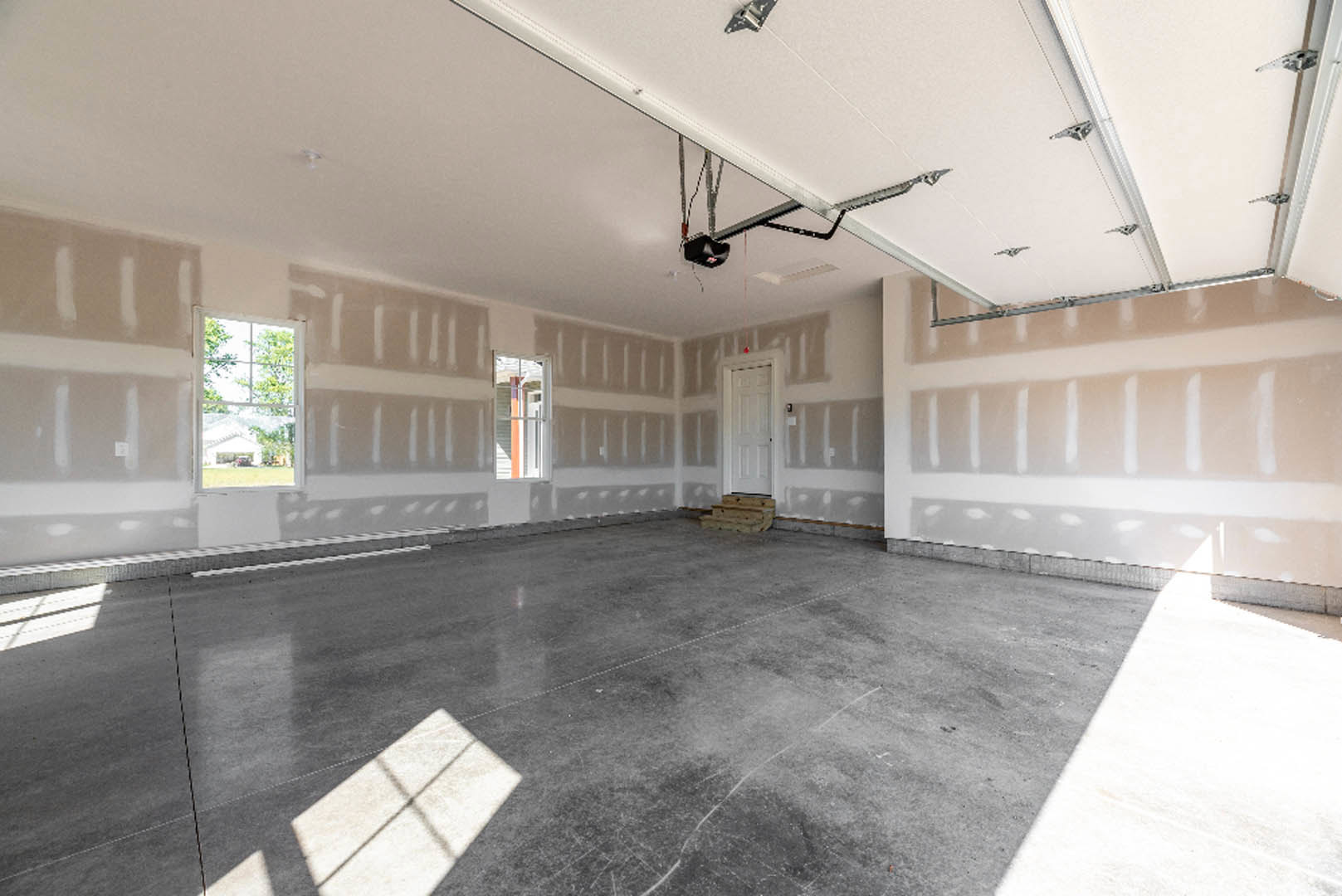 Concrete floor with scattered pallets, white plaster walls, white door, ceiling light fixture, staircase holding stacked wood, black object with illuminated feature, window