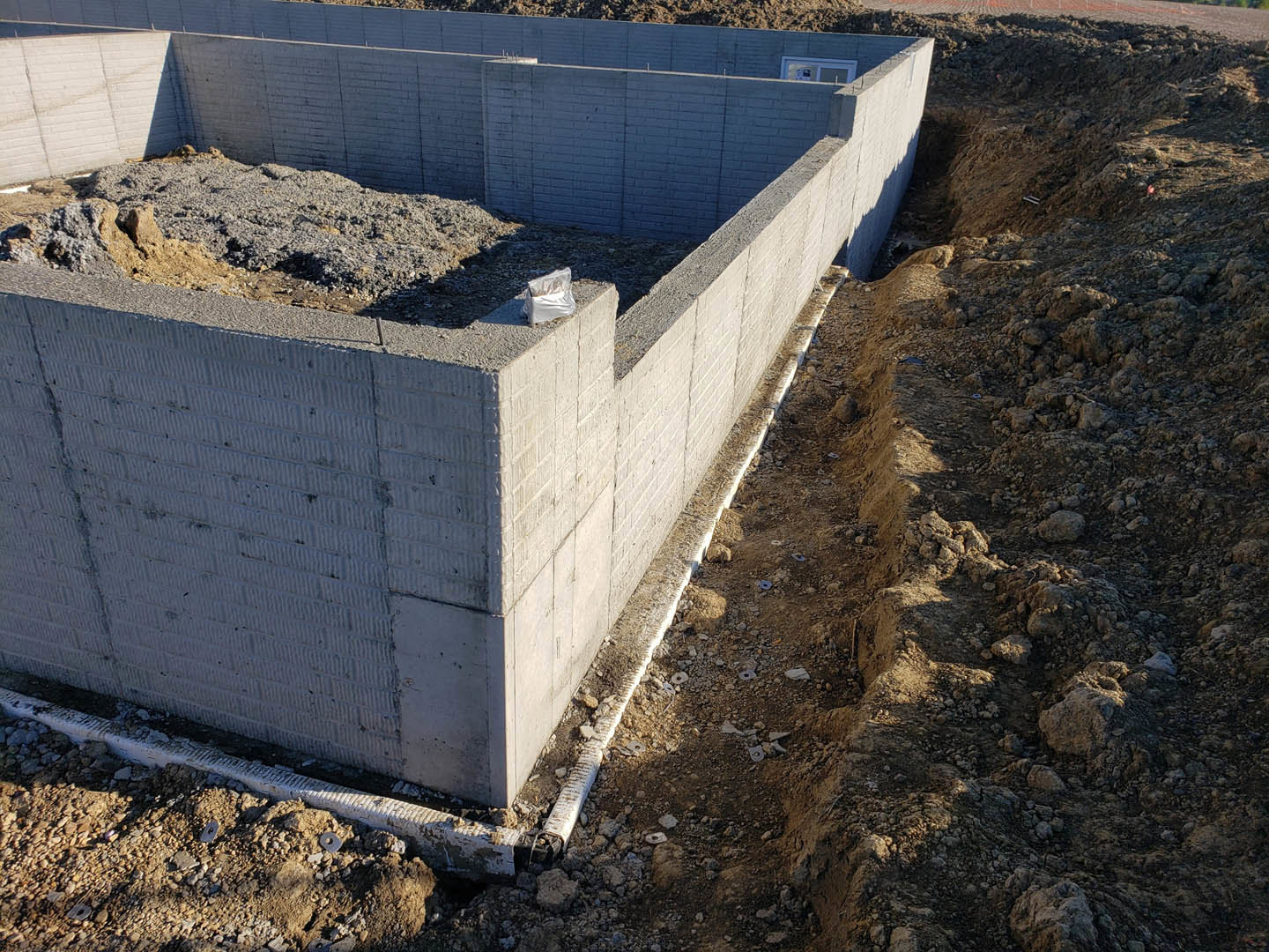 Concrete foundation under construction with exposed soil, drainage channel, unfinished wall, white bag on ground, and scattered rocks