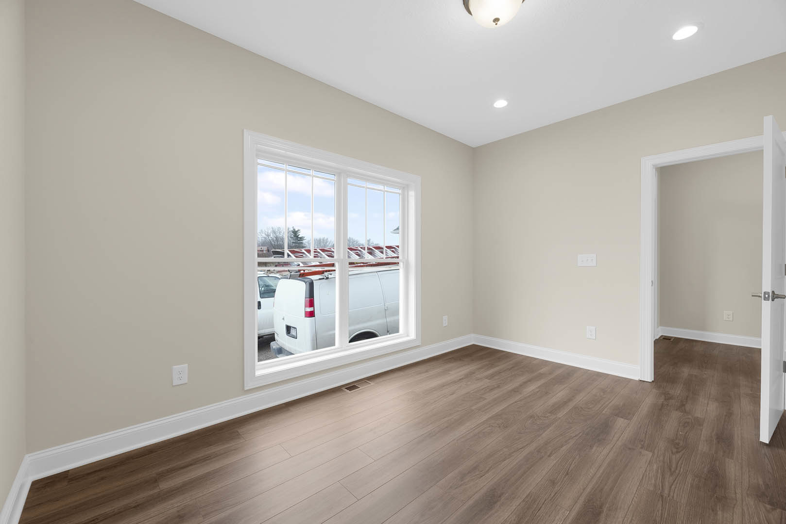 Hardwood floor room with open door, large window overlooking parking lot, ceiling light fixture, plaster walls