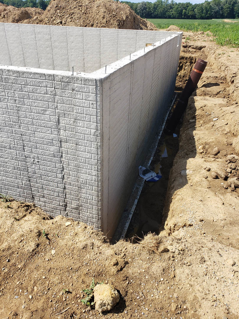 Concrete foundation wall with exposed pipe emerging from soil, rough ground surface, sparse plants, and distant trees under open sky