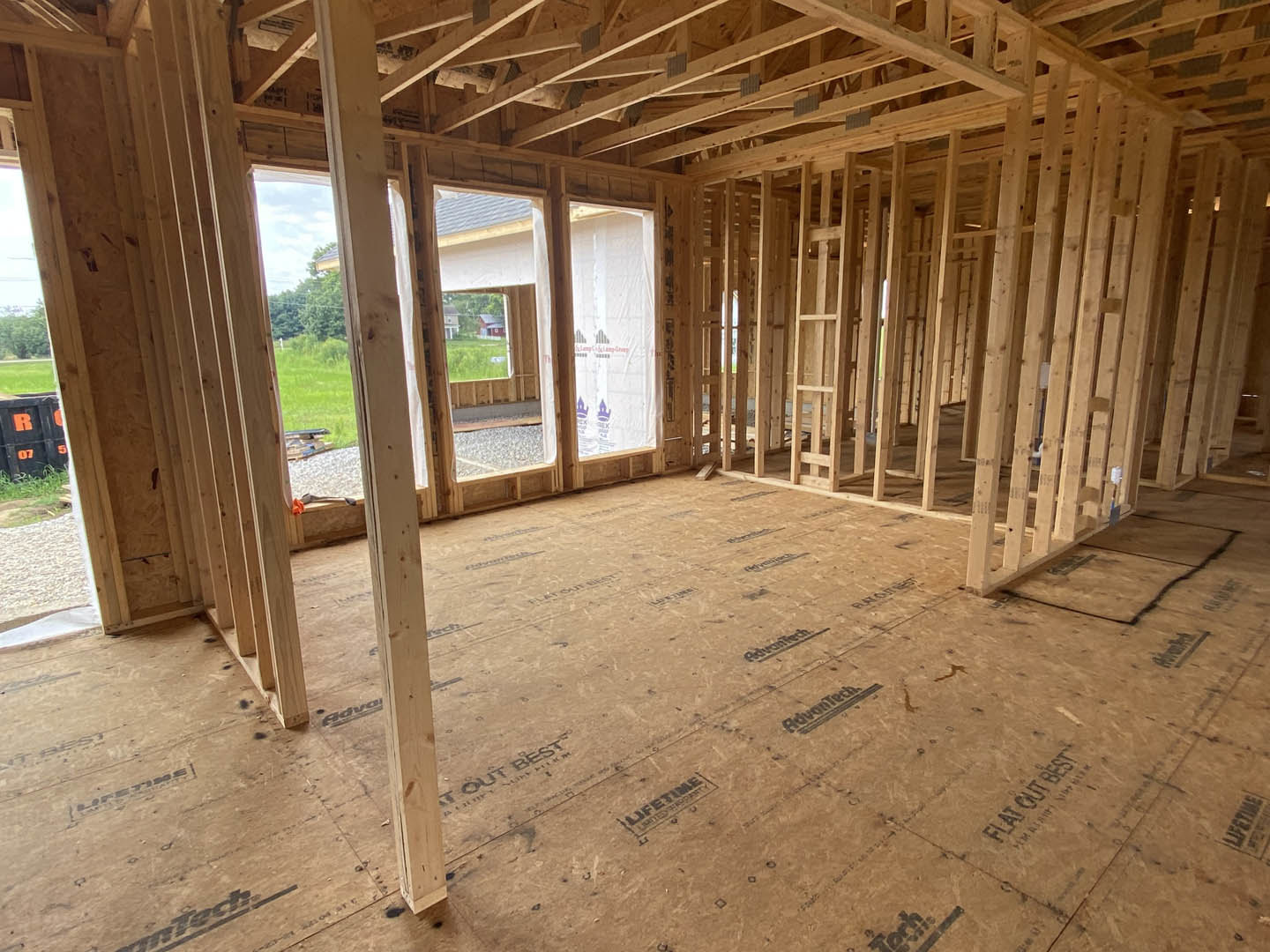 Wood-framed room under construction with exposed beams, plywood floor marked with black text, black container labeled in orange, white plastic sheeting with red lettering, and