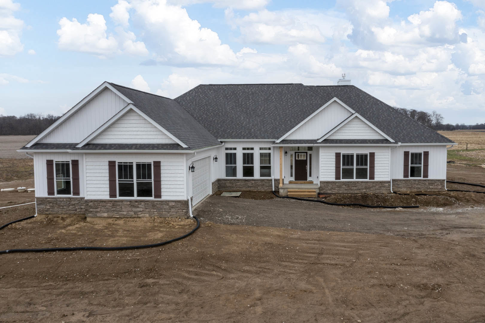 Partially built house with exposed framing, white-trimmed window, front porch, dirt driveway, garden hose, and cloudy sky above