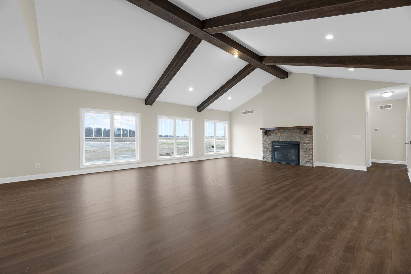 Spacious living area with hardwood floors, exposed wooden ceiling beams, plaster walls, large glass windows overlooking a field, and a fireplace with a wood mantle
