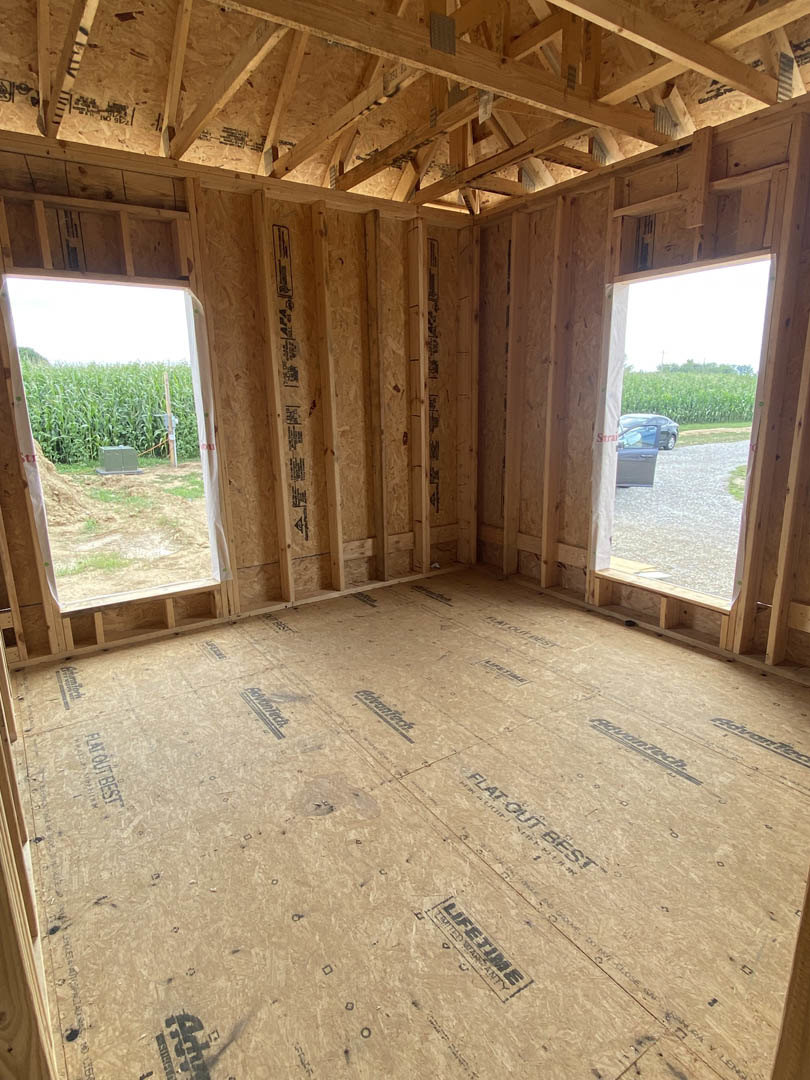 Sunlit room featuring large windows, finished wood plank flooring, exposed ceiling beams, and partially installed wall insulation