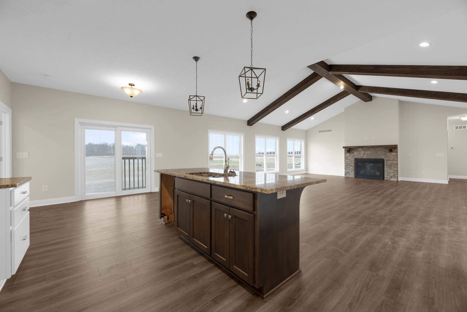 Spacious open floor plan featuring hardwood floors, marble kitchen island with sink, modern ceiling light fixture, stone fireplace wall, sliding glass door with metal railing, and