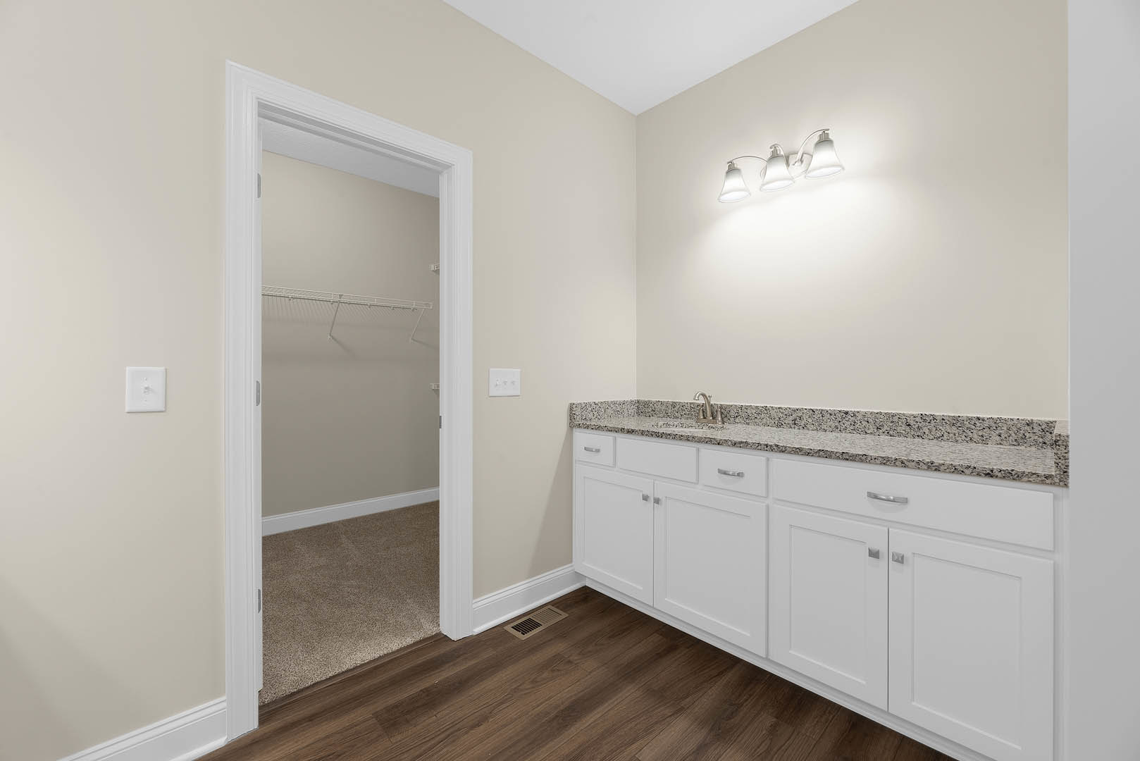 Bathroom with white cabinets, wood flooring, white light switch on wall, row of white lights above mirror, closet with white door, floor vent, granite countertops on cabinetry