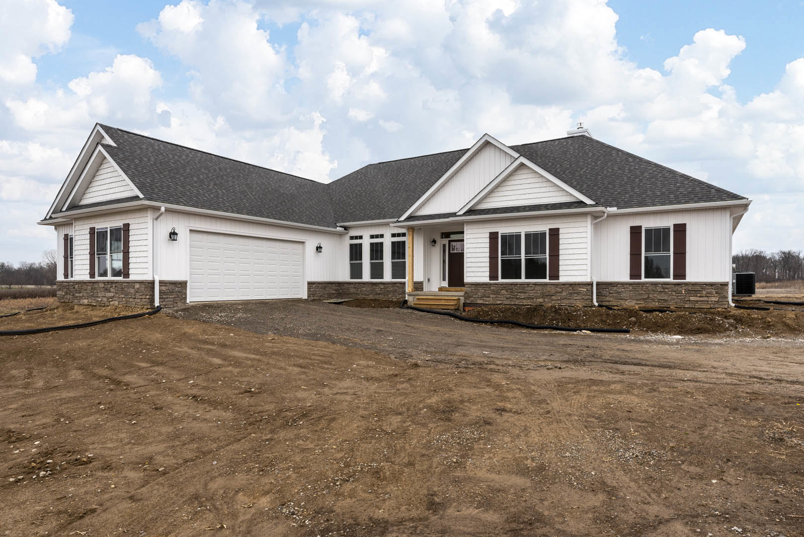 Two-story house with white-trimmed windows, attached garage, and front door facing a dirt road; cloudy sky overhead, black square object with white label near the driveway.