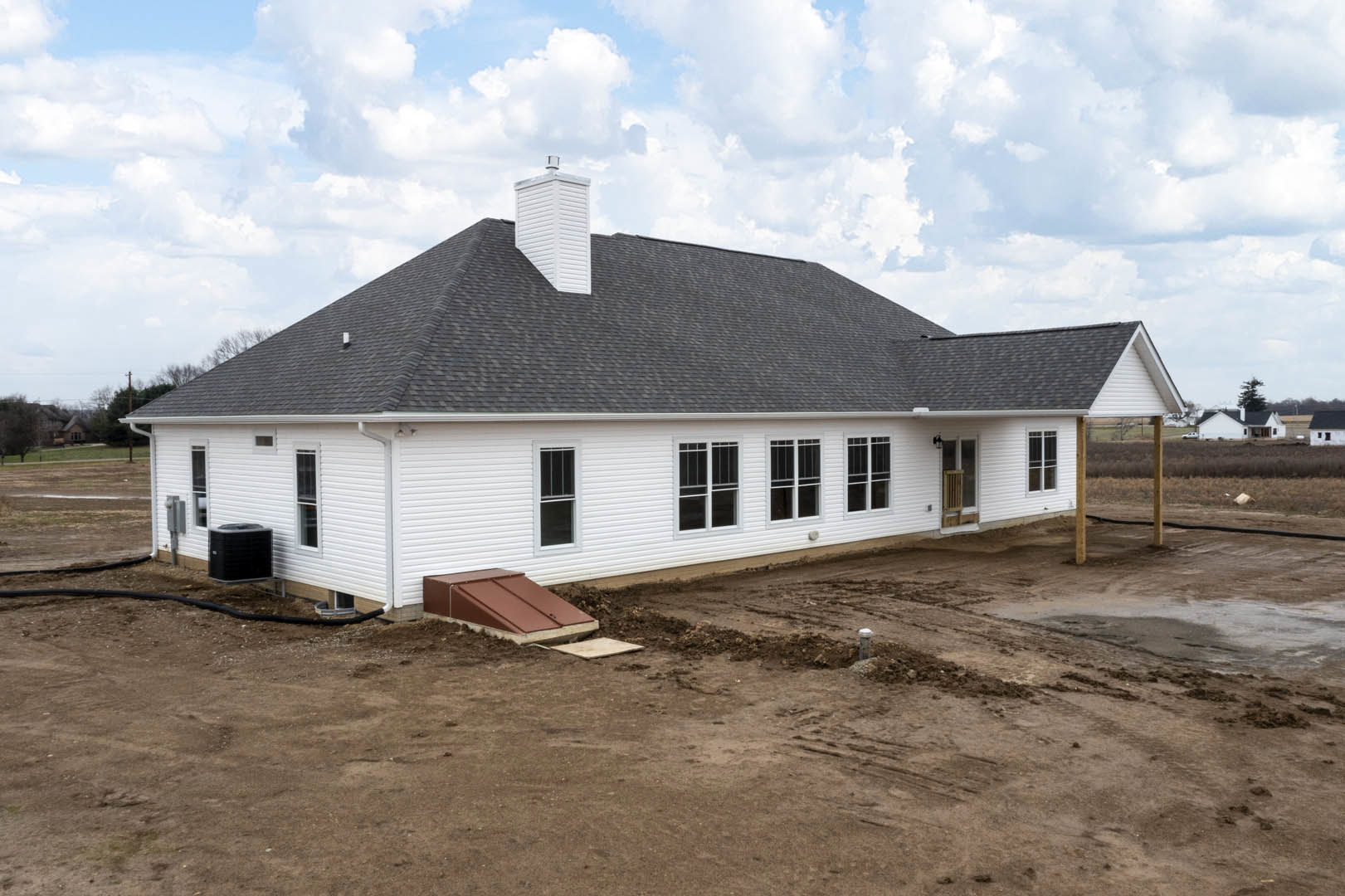 White house with brown roof and chimney, black vent on side wall, dirt yard in foreground, windows along exterior, cloudy sky overhead