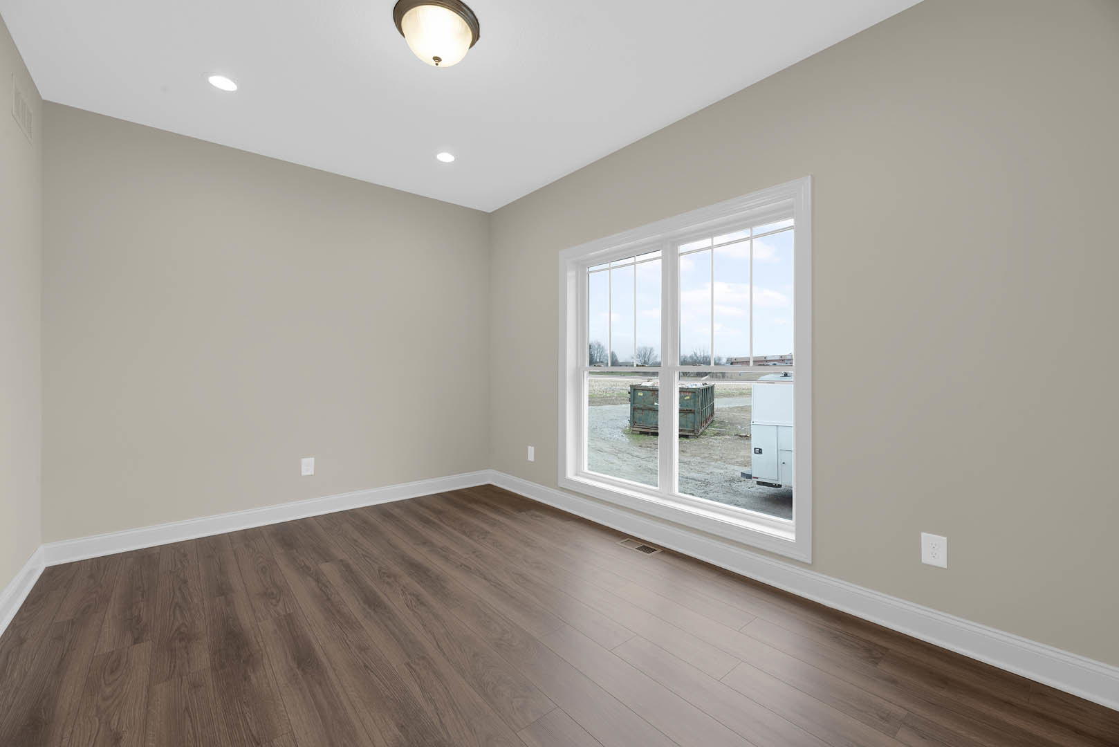 Sunlit room featuring wide-plank hardwood flooring, large window overlooking green yard, white walls, and recessed ceiling light.