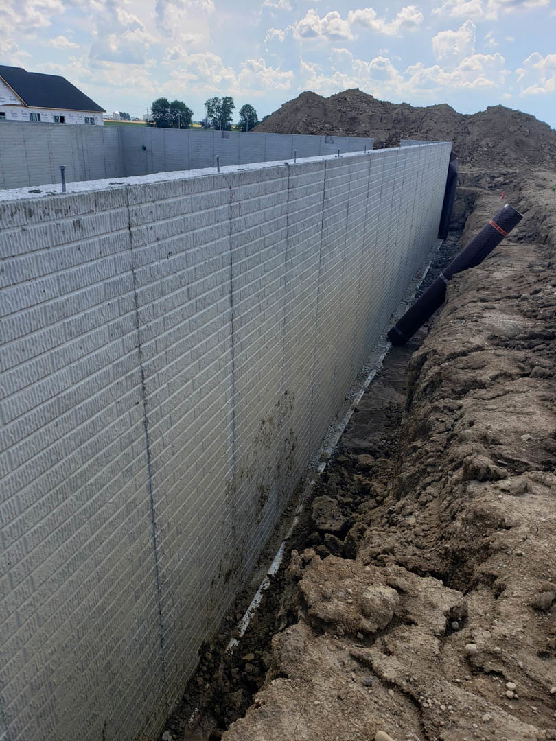 White exterior wall with a vertical pipe running through the center, brown dirt ground, sloped roof, blue sky with scattered clouds, distant mountain, and a tree in a grassy field