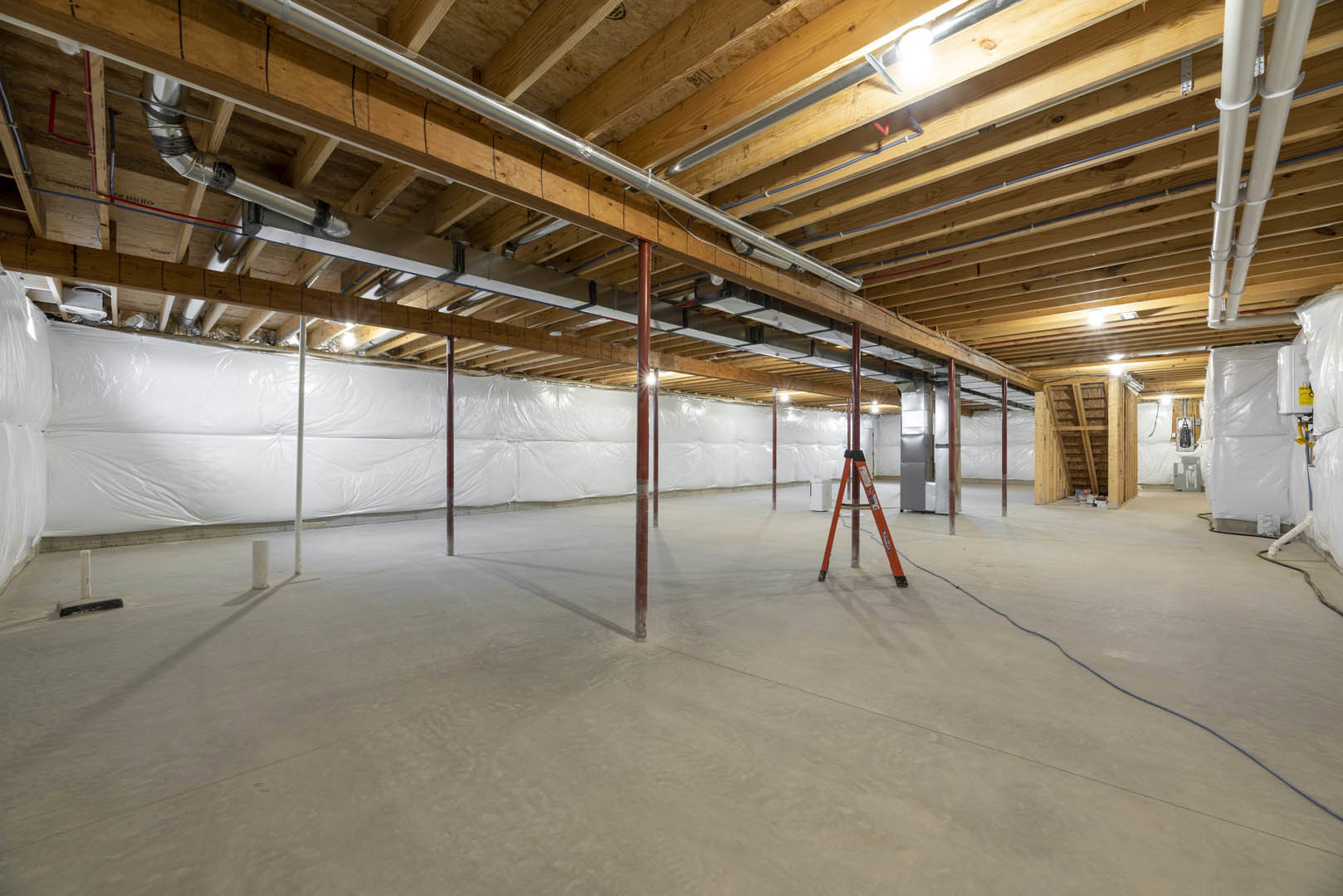 Open room with white walls, exposed wooden ceiling beams, metal pipes, concrete floor, and a vertical ladder attached to a pole.