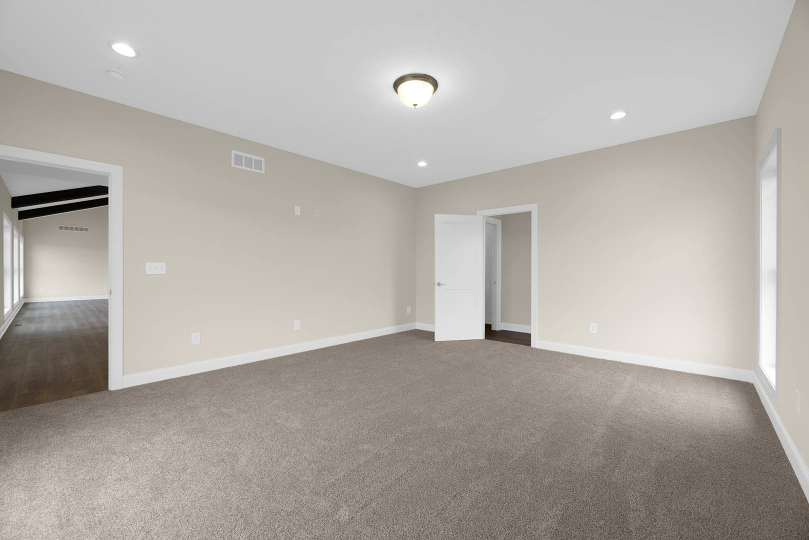 Carpeted room with white walls, open white door with silver handle, ceiling light fixture, adjacent room with wood flooring