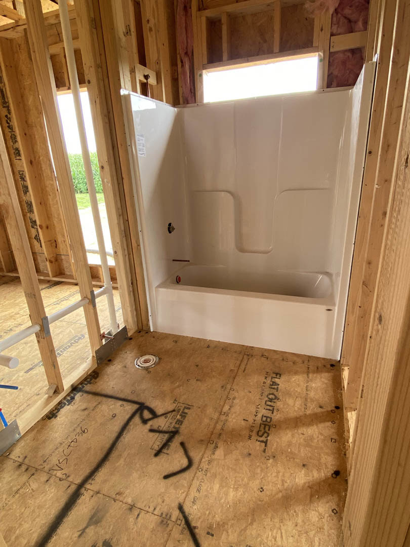 Freestanding white bathtub set in a wood frame, wood plank flooring with black script, plaster walls, large window, modern light fixture