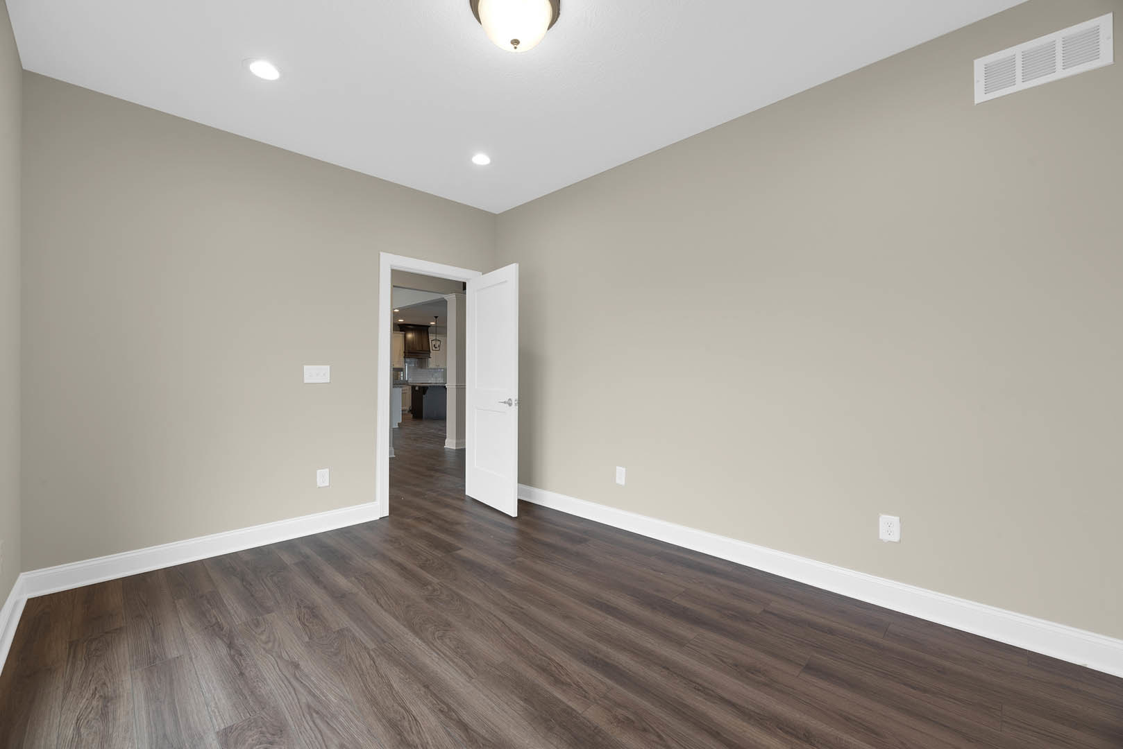 Dark wood laminate flooring, white door with silver handle open to adjacent room, ceiling vent and recessed light, white walls with crown molding.