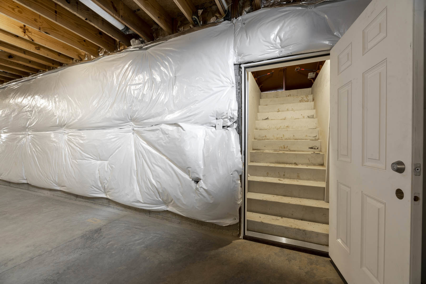Wood staircase with plastic covering leads down to unfinished basement with white plaster walls, white door with silver knob, exposed pipe, and concrete floor.