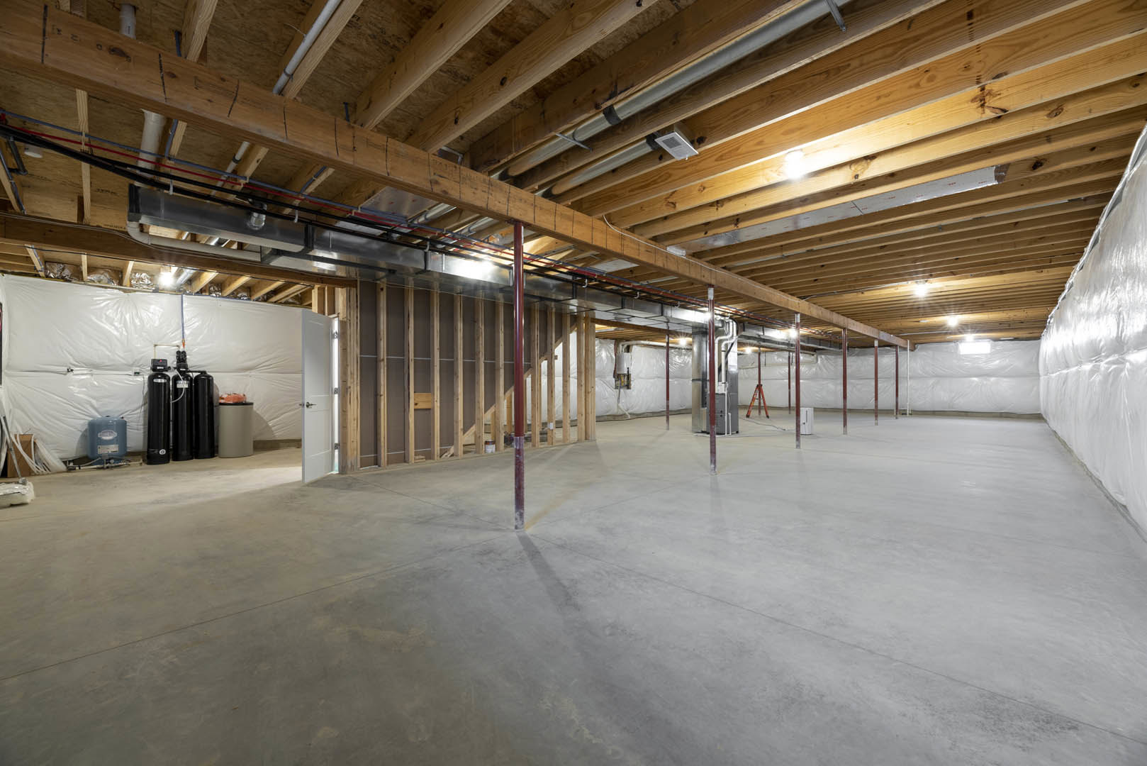 Spacious unfinished basement with exposed wood ceiling beams, concrete floor, black water tanks, white cylinder with black top, metal support pole, and blue storage container