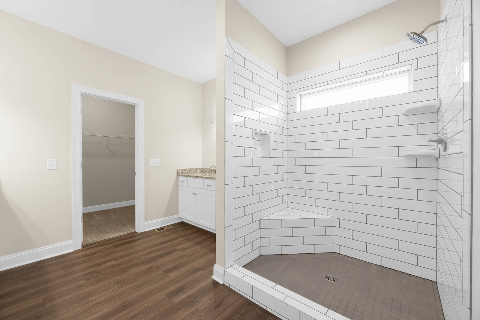 Bathroom with wood flooring, tiled shower enclosure, metal shower head and pole, wall-mounted light fixture, white window frame and shelf, plaster walls, and shower drain.