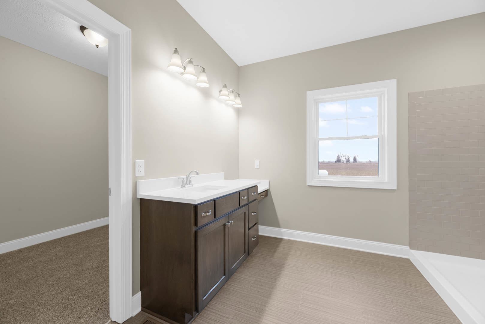 Bathroom with white vanity and undermount sink, three-light fixture above mirror, window overlooking grassy field, white tile flooring, partial view of carpeted area