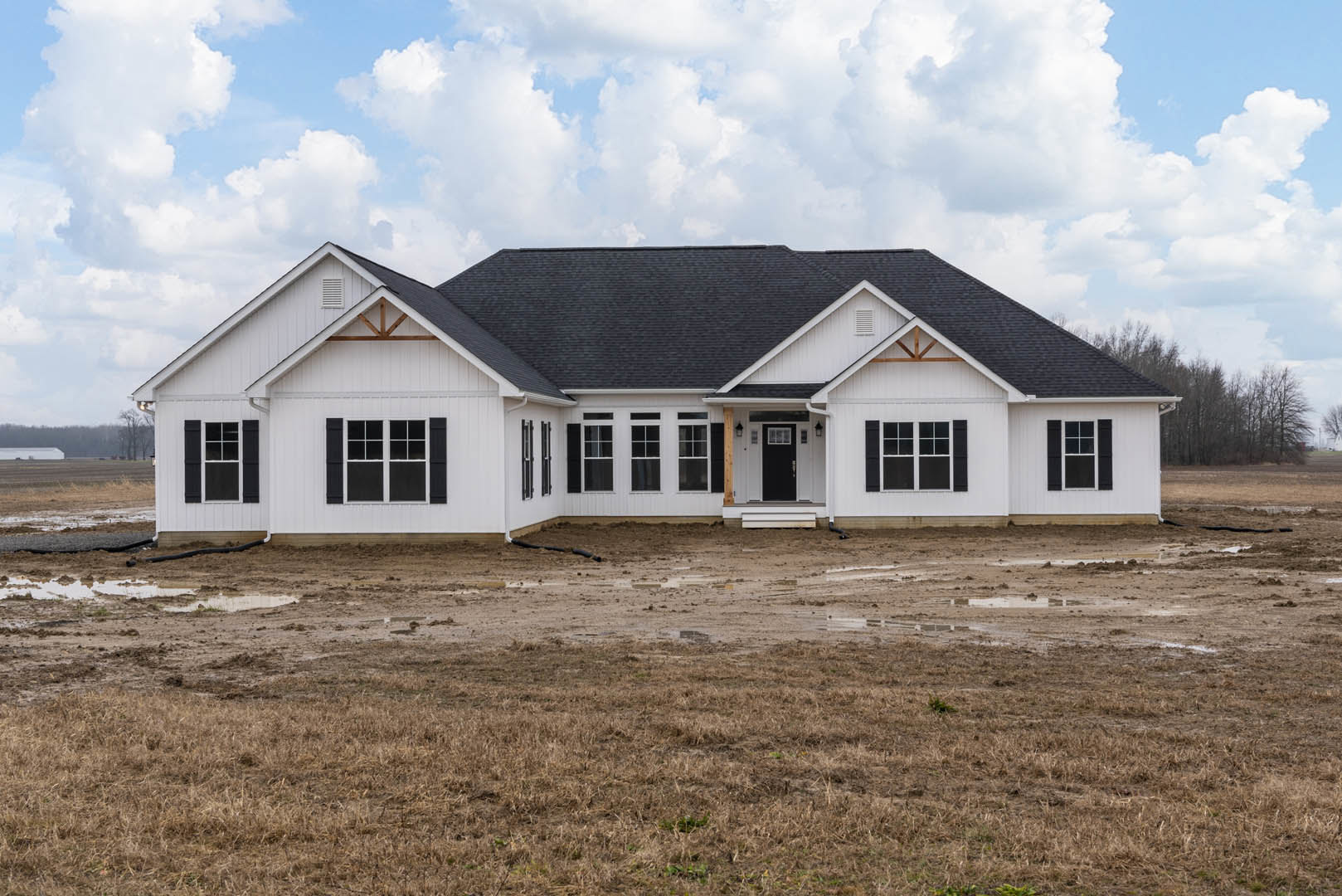 White house with black roof, multiple windows, and black door, set beside a dirt field with a garden hose under a cloudy sky
