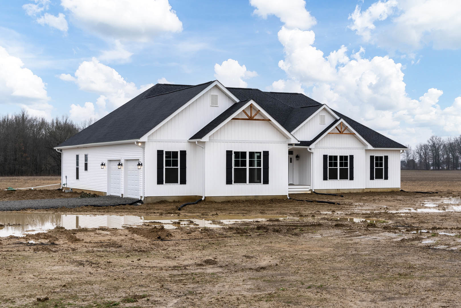 White siding house with black roof and black shutters, multiple windows, muddy yard with exposed pipe, cloudy sky overhead