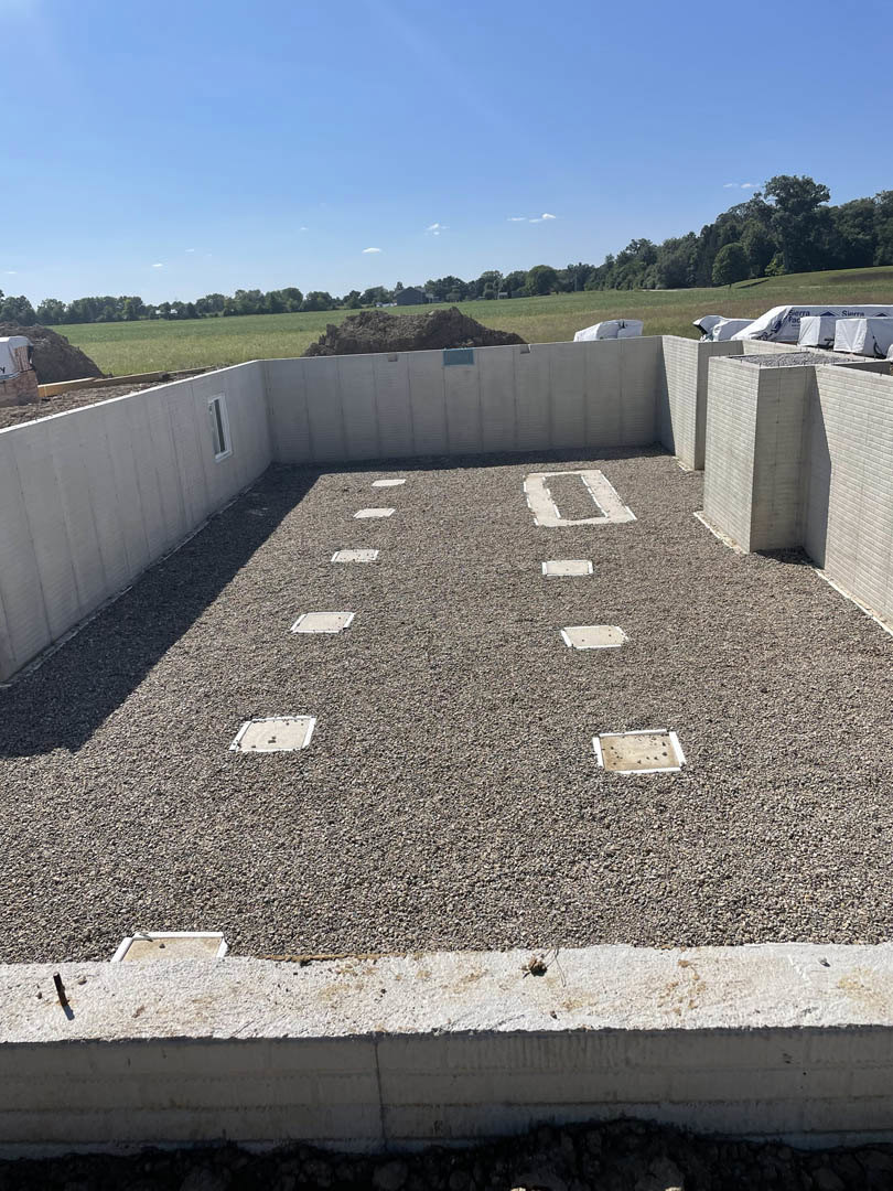 Concrete foundation with square block openings, gravel ground, person standing nearby, blue sky with scattered clouds, tree in background
