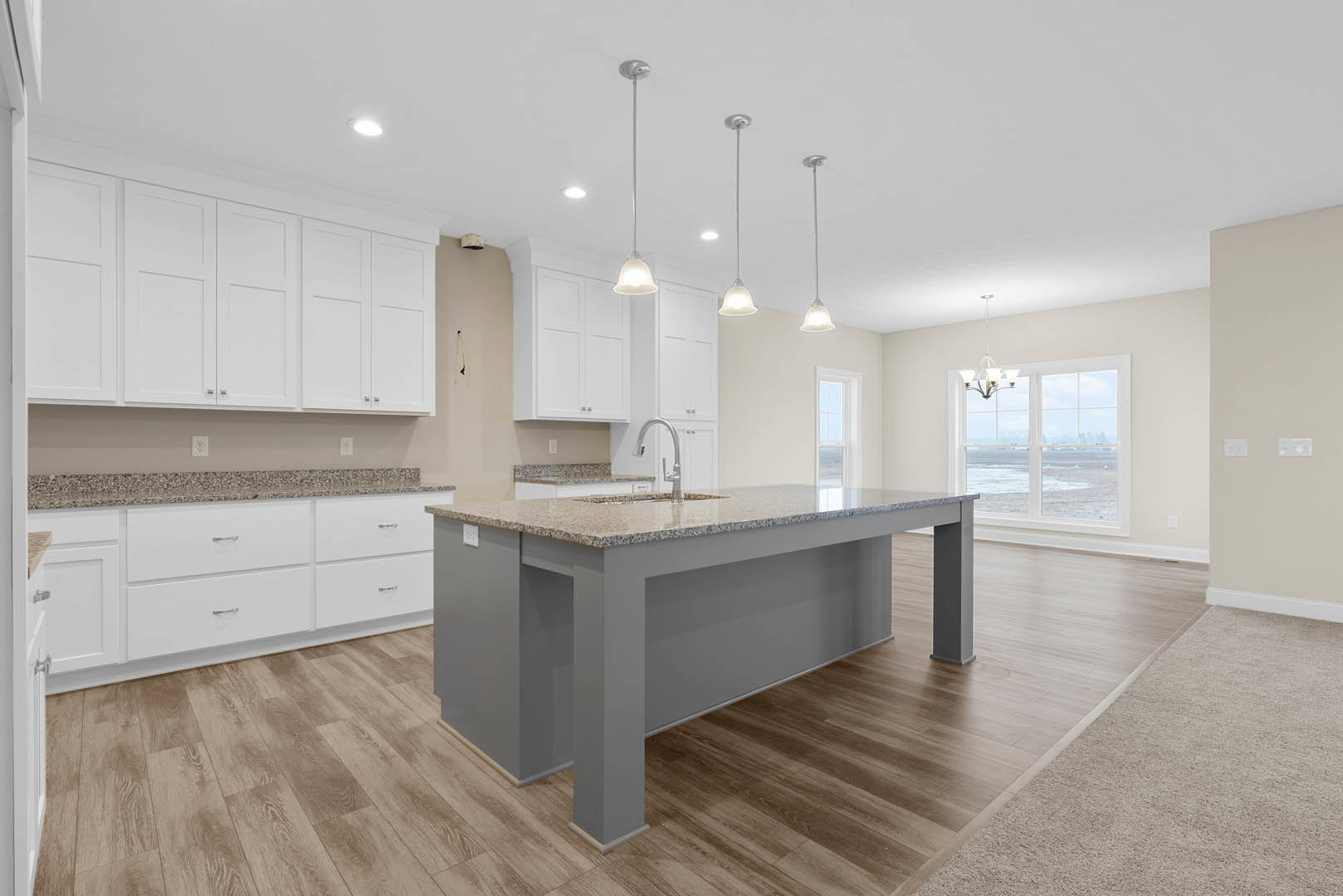 Kitchen with central island featuring built-in sink, white cabinets with silver knobs, wood flooring, granite countertops, large window, and modern pendant light fixture.