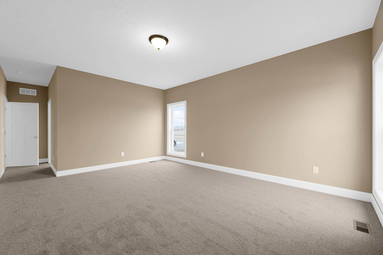 Beige-walled room with white trim, carpeted floor, large window showing outdoor view, white door with silver handle, ceiling-mounted light fixture