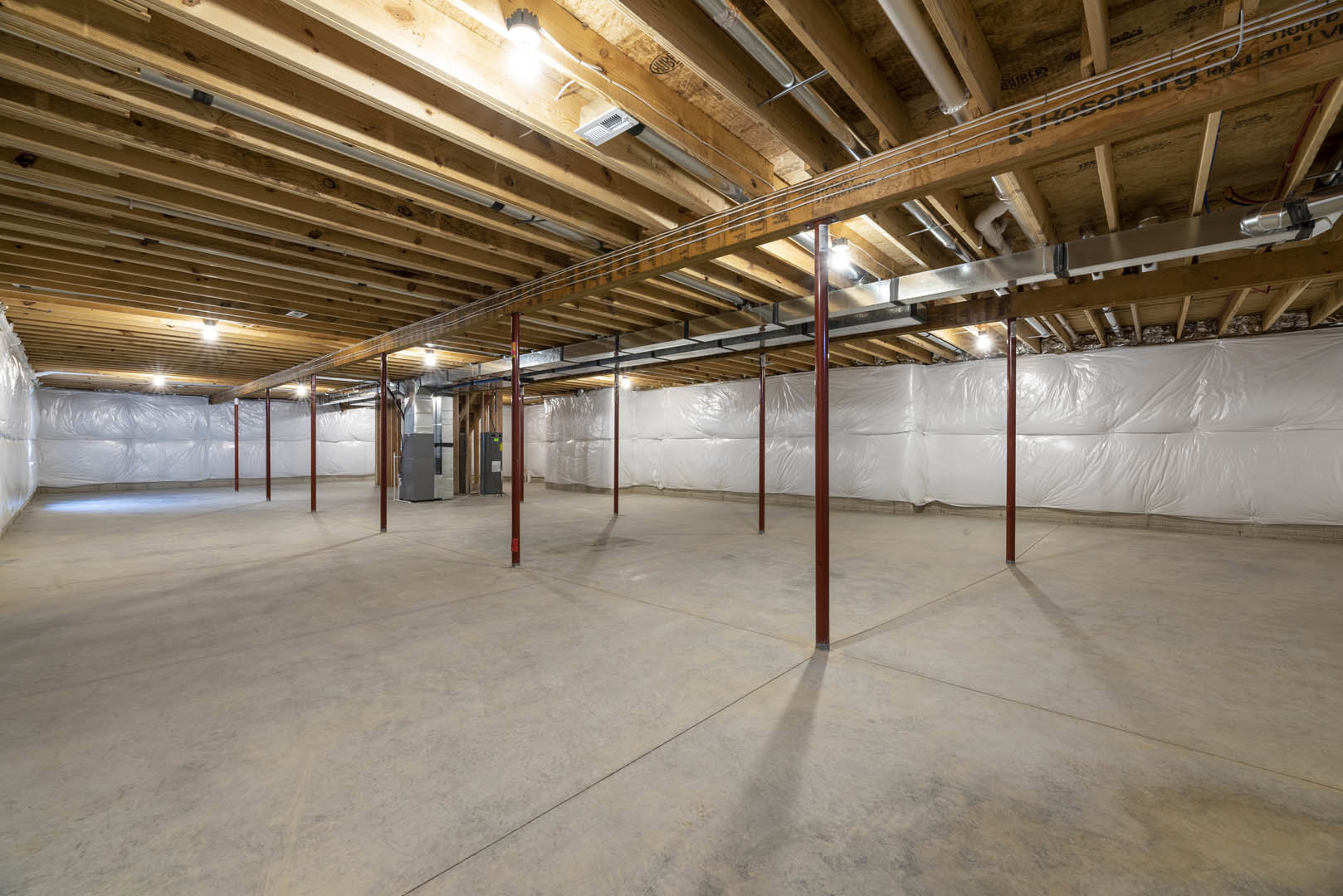 Exposed wooden ceiling beams, concrete floor with red support poles, large white bag, grey rectangular object with brown frame, unfinished basement walls and visible pipes