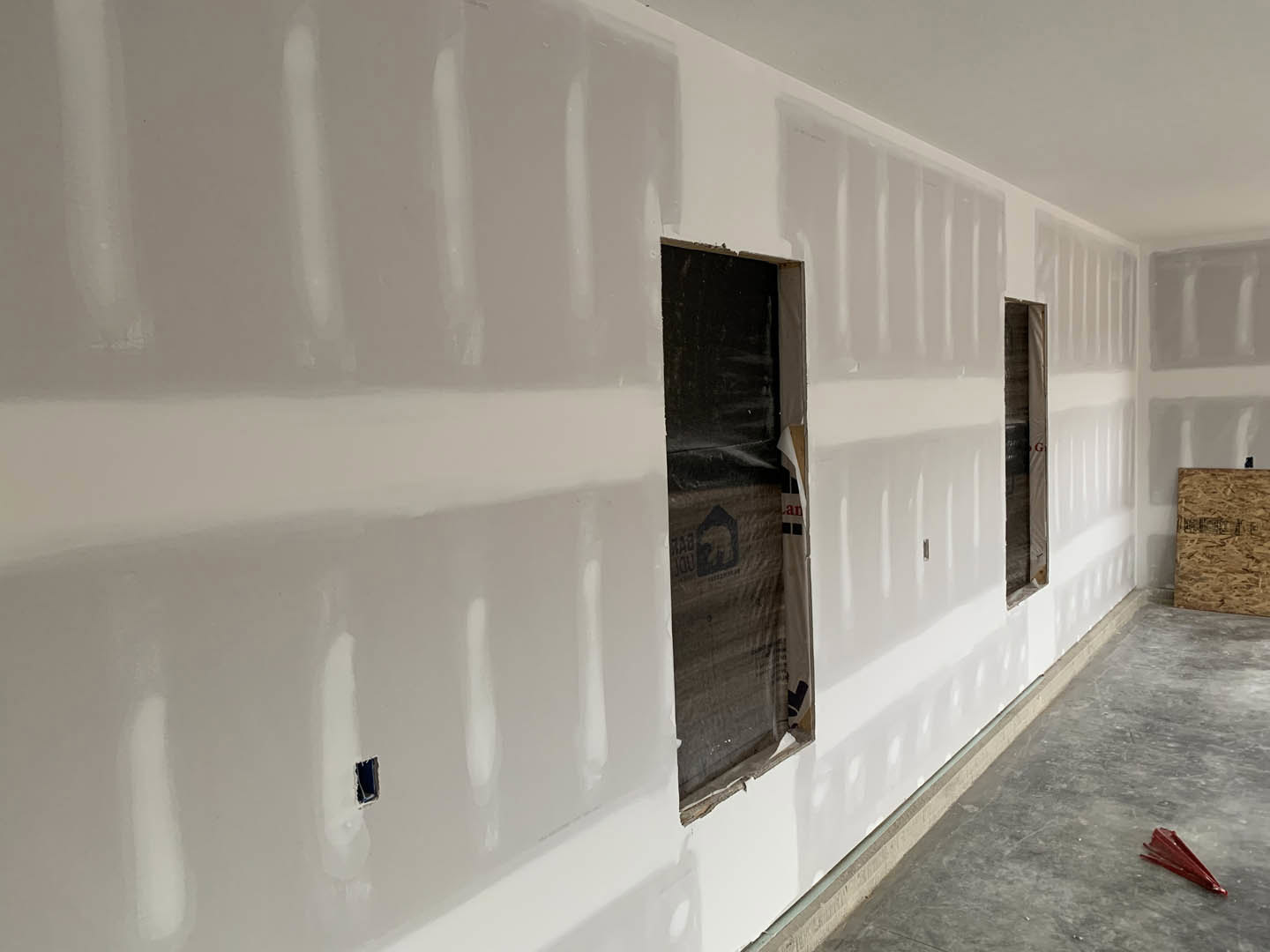 Hallway with plaster walls, multiple rectangular windows, wood panel detail, red tool resting on grey floor, plastic bag with logo near wall