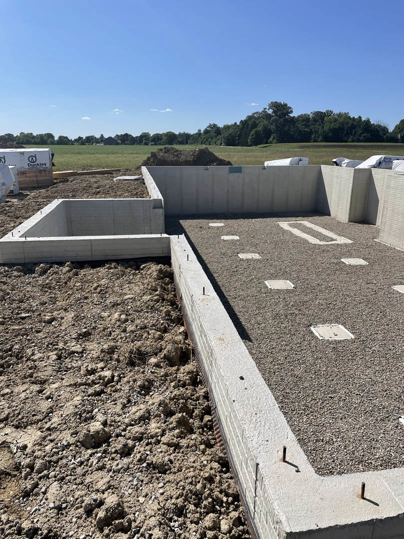 Concrete foundation and stone retaining wall surrounded by dirt and gravel, white tarp with black text, blue sky with scattered clouds