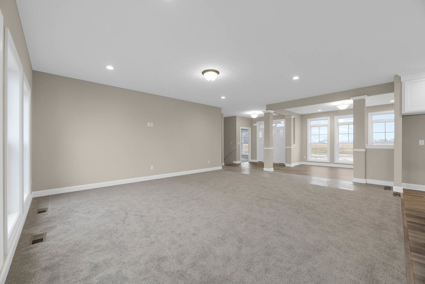 Spacious carpeted room with tall white wall, ceiling vent, recessed light fixture, staircase with wood railing, and windows showing blue sky