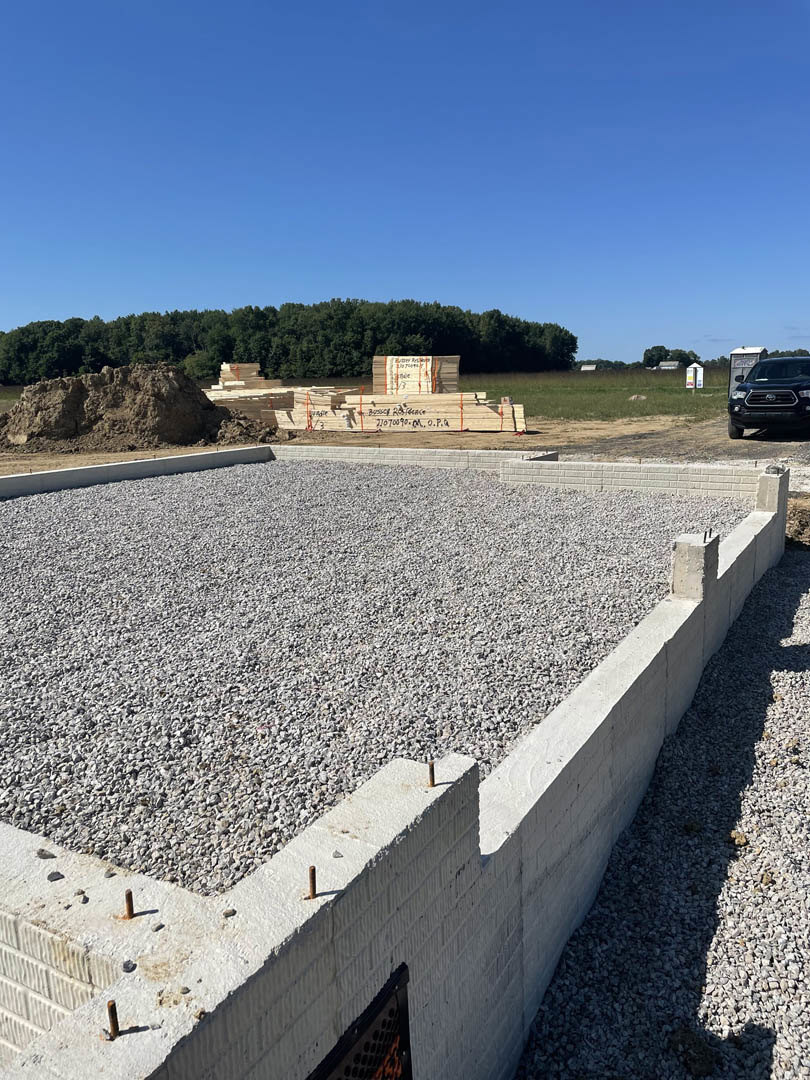 Black car parked beside newly poured concrete foundation with gravel, portable toilet nearby, group of trees in background under clear blue sky, close-up of rocks in foreground