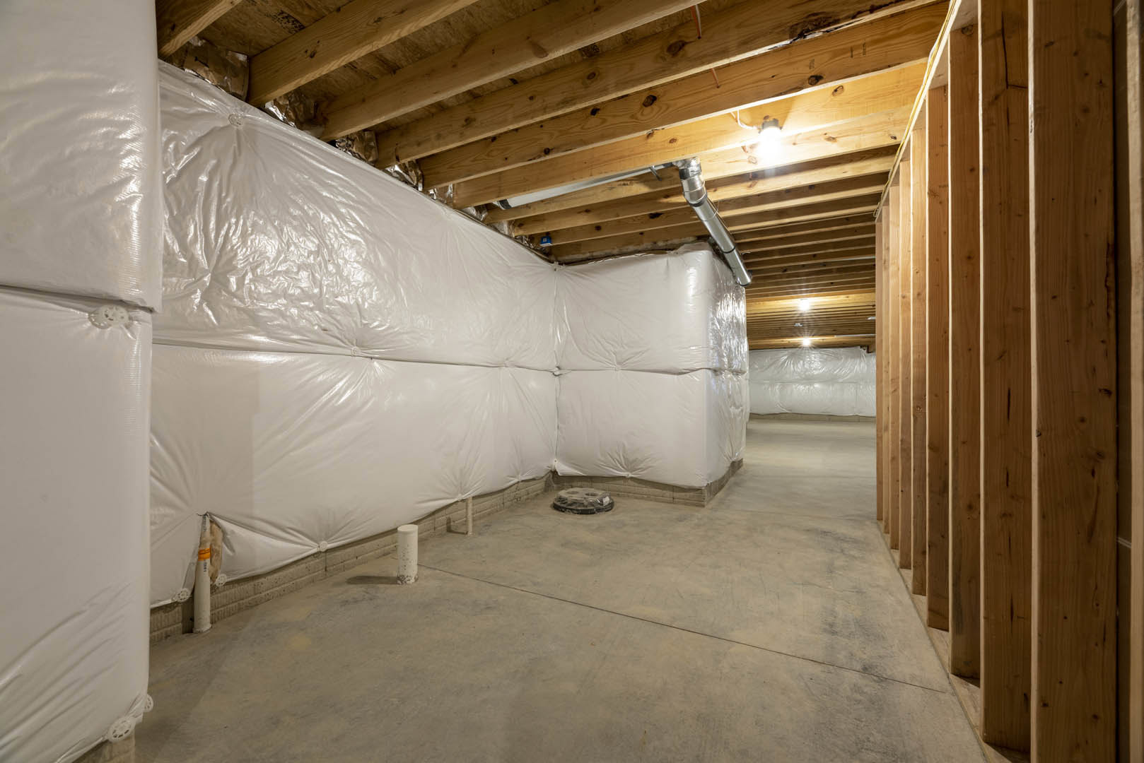Room under construction with white plastic sheeting covering walls, exposed wooden ceiling beams, visible pipes, concrete floor, and a white support pole.