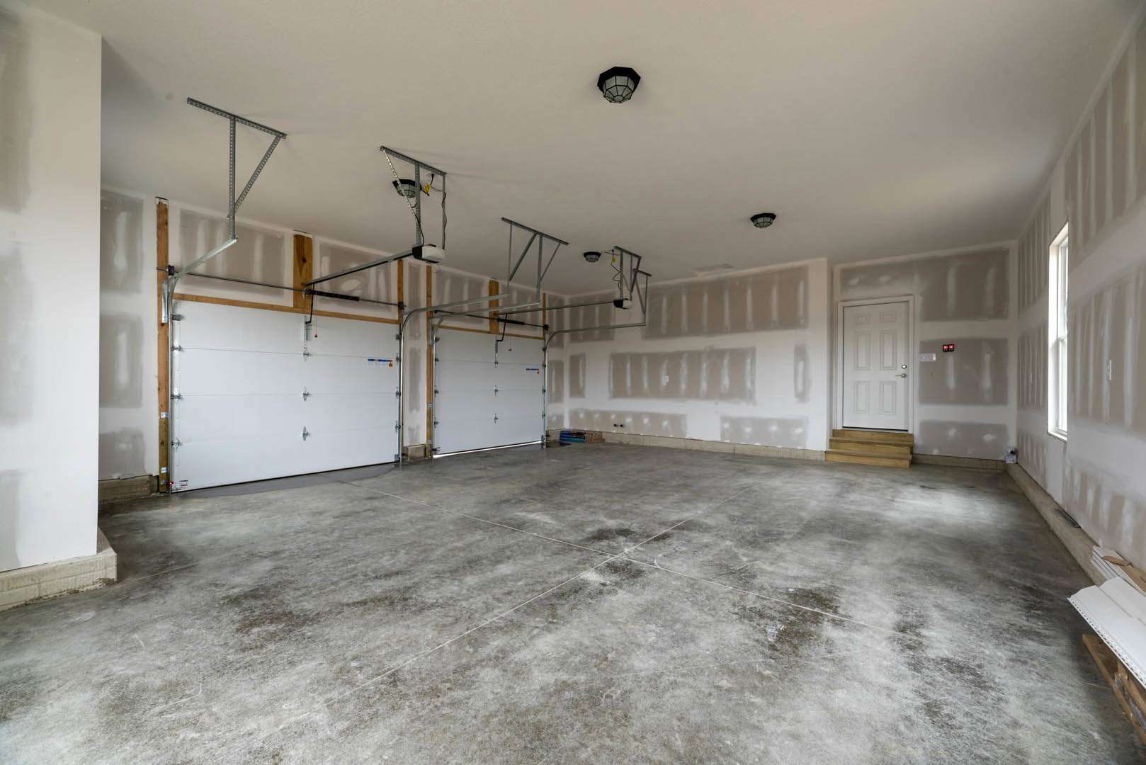Spacious empty room with smooth white plaster walls, polished concrete floor featuring subtle white lines, white door with silver handle, ceiling-mounted light fixture, and garage