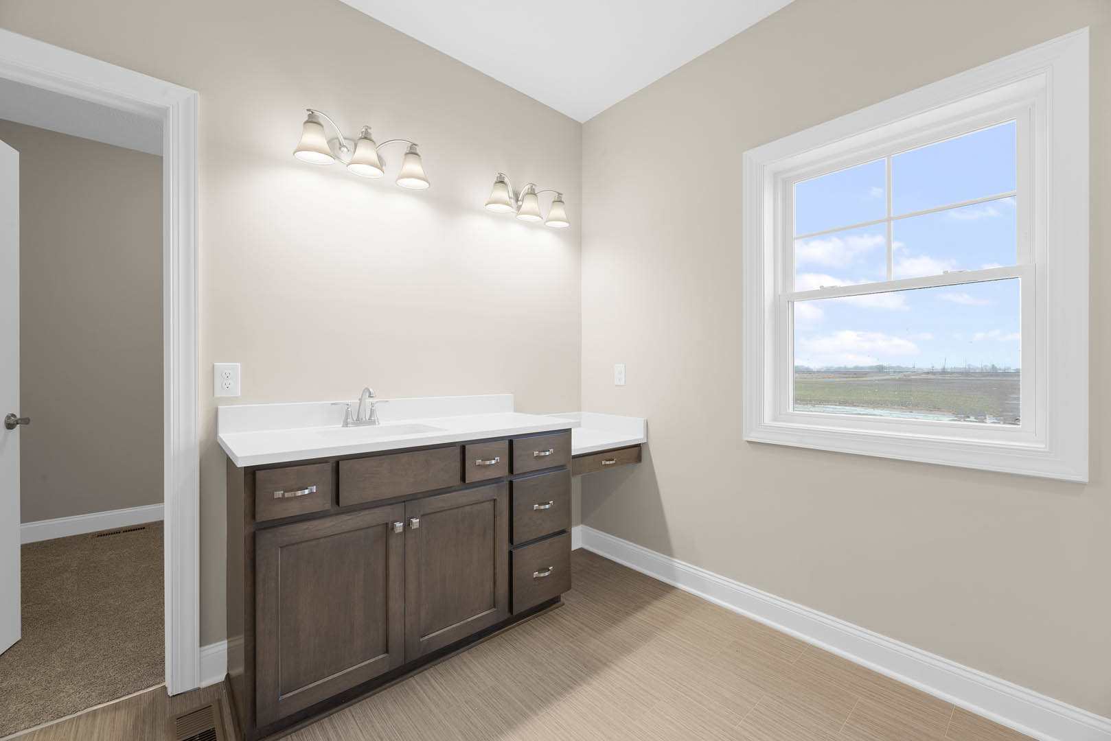 Bathroom with white tile walls, wood vanity with integrated sink, chrome faucet, and a window overlooking a field and blue sky; overhead recessed lighting and cabinetry drawers