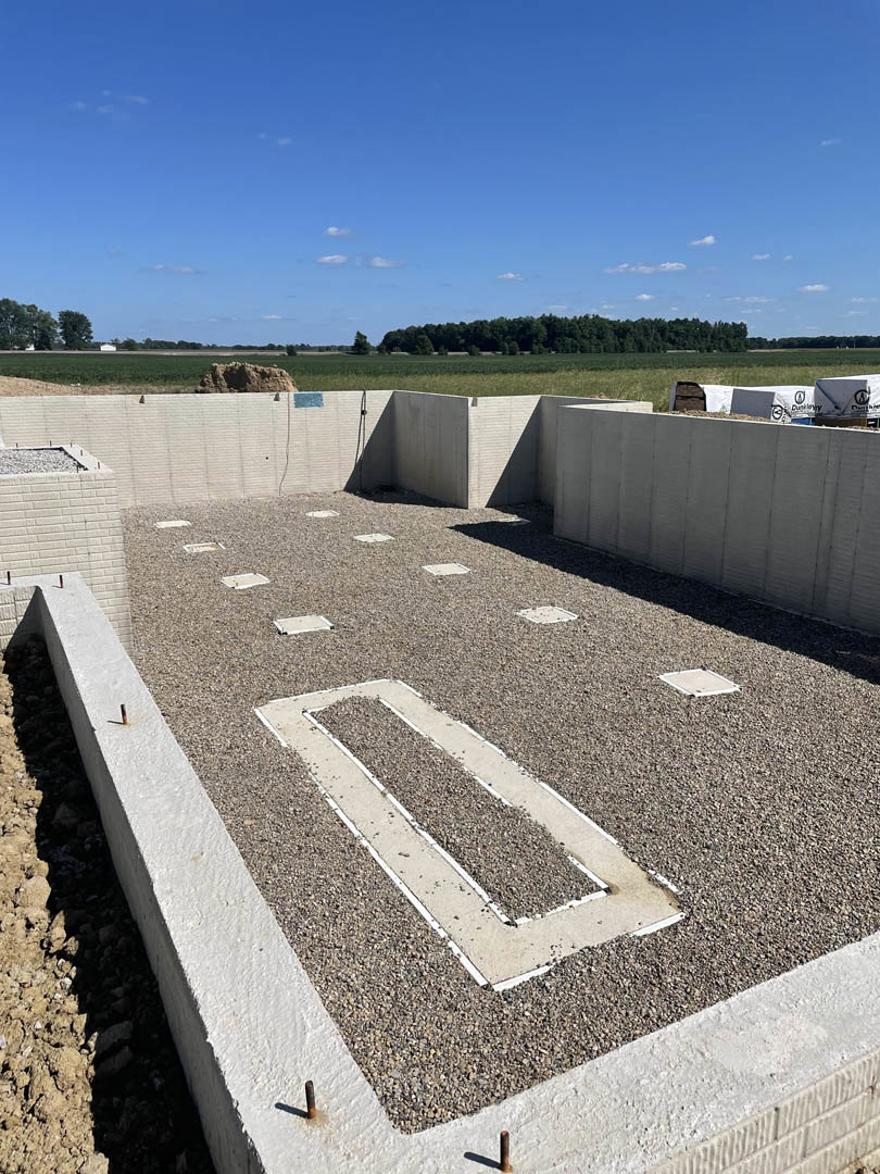 Gravel building site bordered by a large grassy field and trees, white rectangular construction material on gravel, gravel road with painted white lines, blue sky with scattered