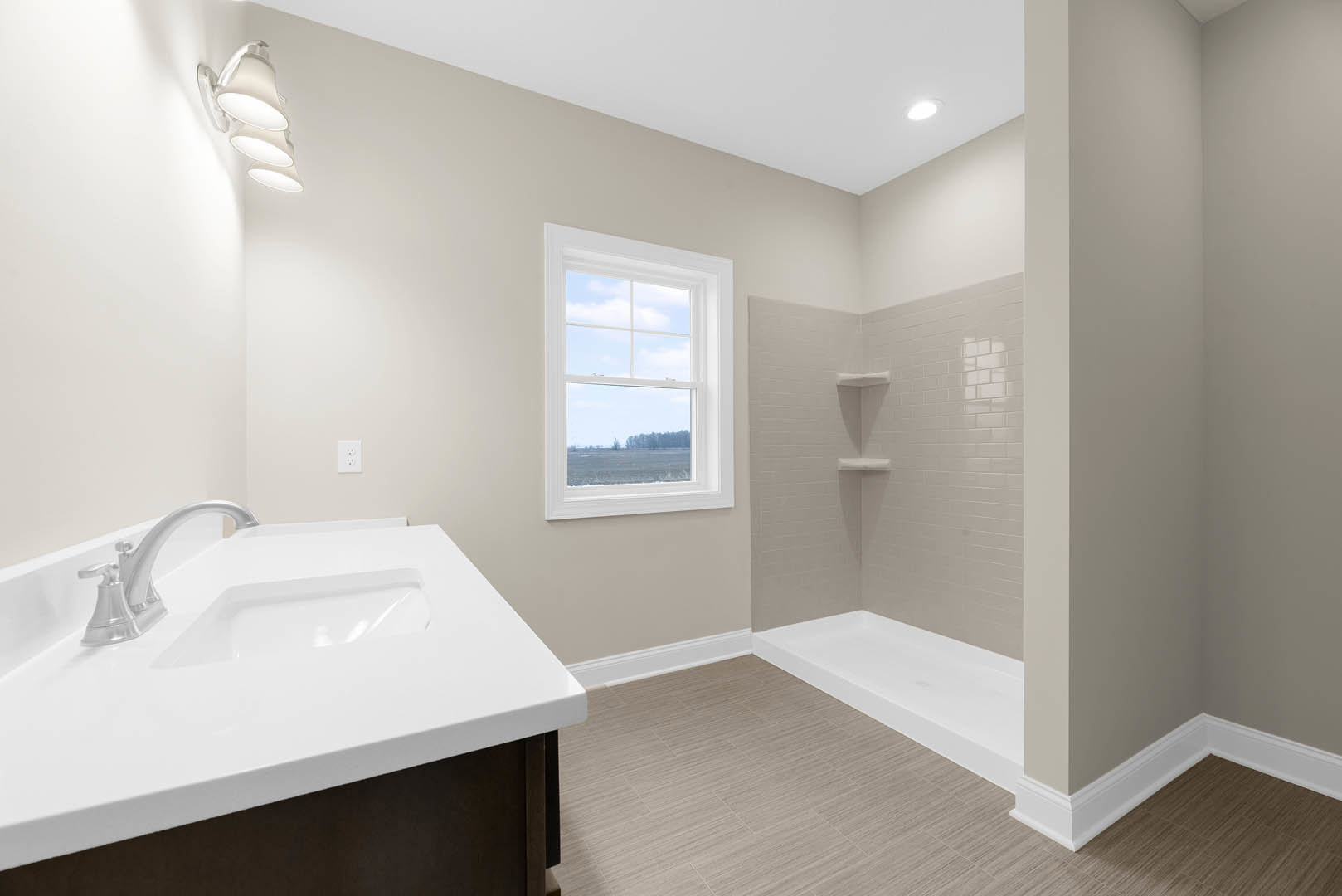 Bathroom with white sink and chrome faucet, glass-enclosed shower, tile floor, window overlooking grassy field, wall-mounted lamp.