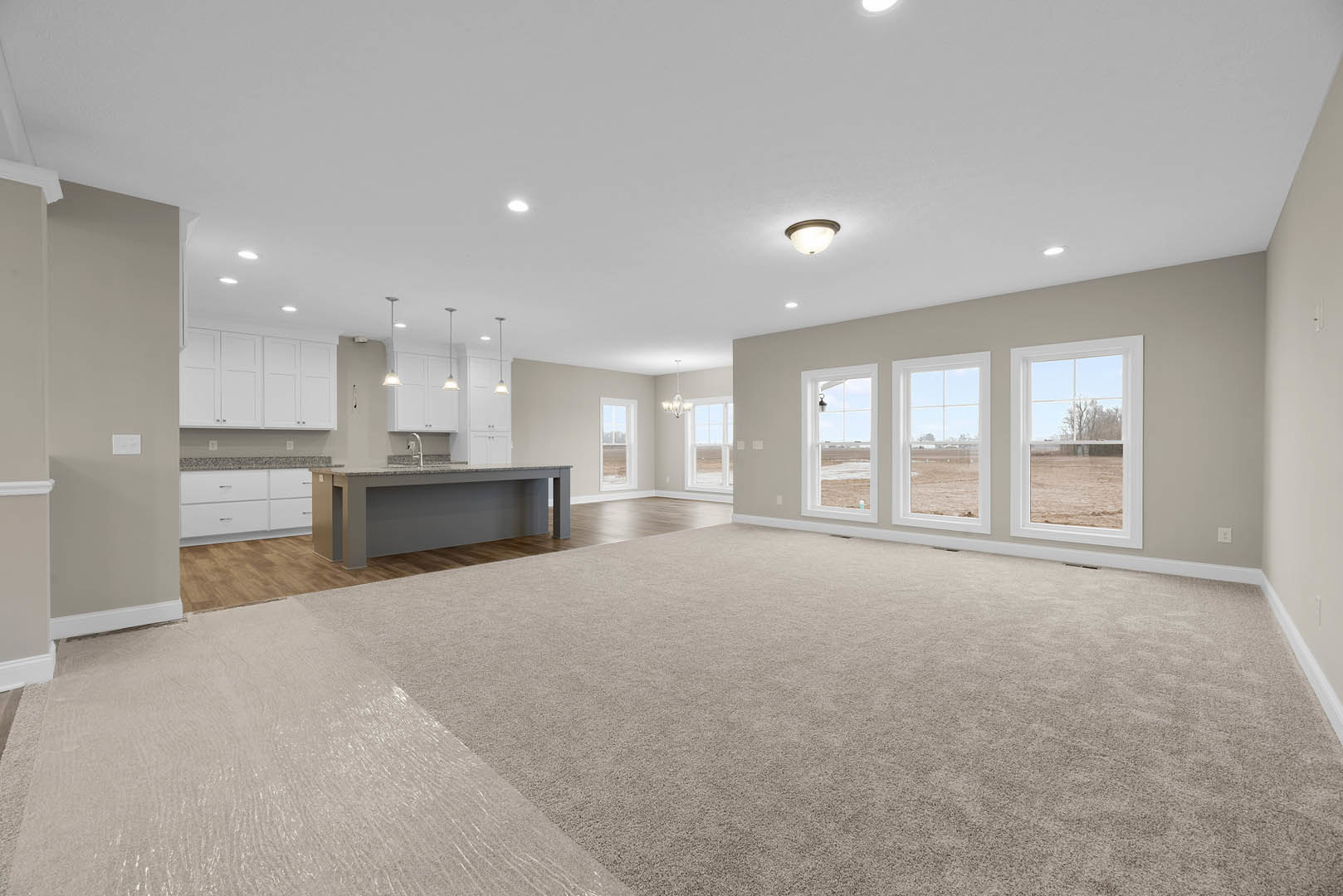 Spacious living room with carpeted floor, open kitchen featuring cabinetry and sink, dining area, white plaster ceiling with recessed lights, large windows overlooking a grassy