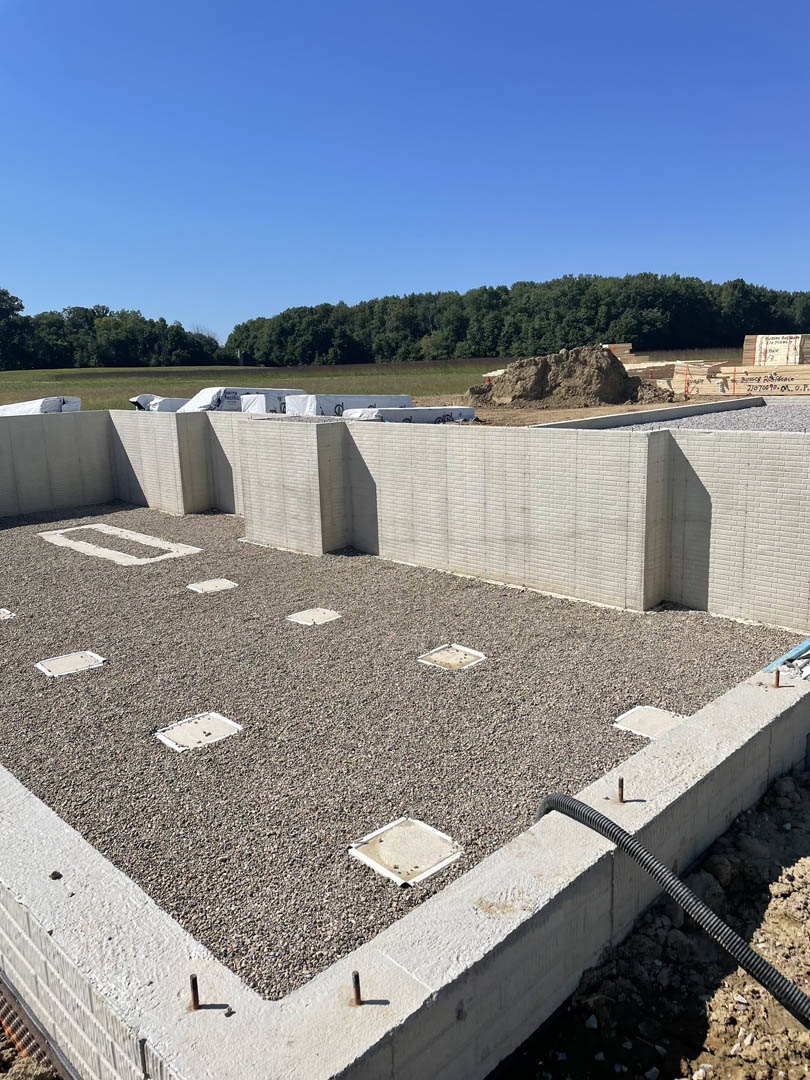 Concrete foundation and partial wall surrounded by gravel, set beside a dense field of trees under a clear blue sky