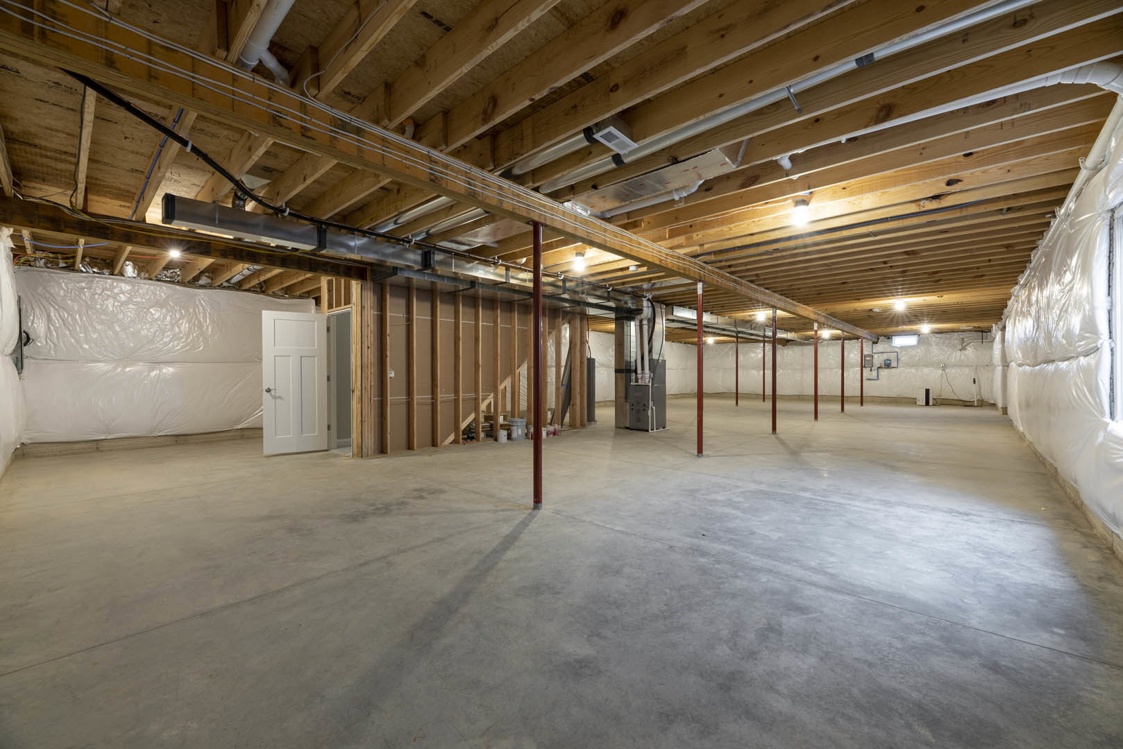 Basement room with exposed wooden beams and ceiling, concrete floor with red steel poles, white door with silver handle, visible pipes, and a white plastic bag hanging on the wall.