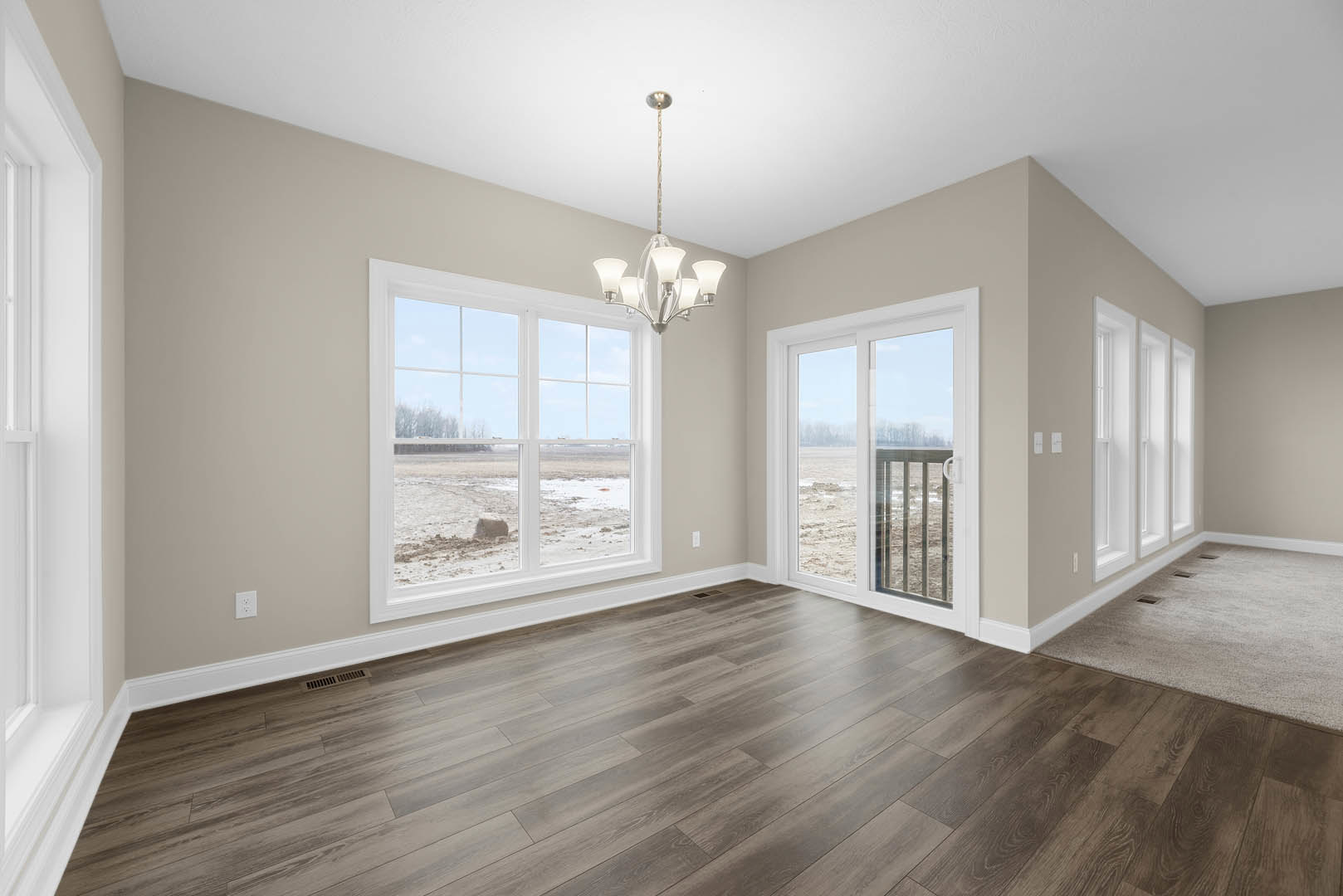 Wood flooring in a spacious room featuring a modern chandelier, large window and sliding glass door overlooking an open field, white walls with crown molding