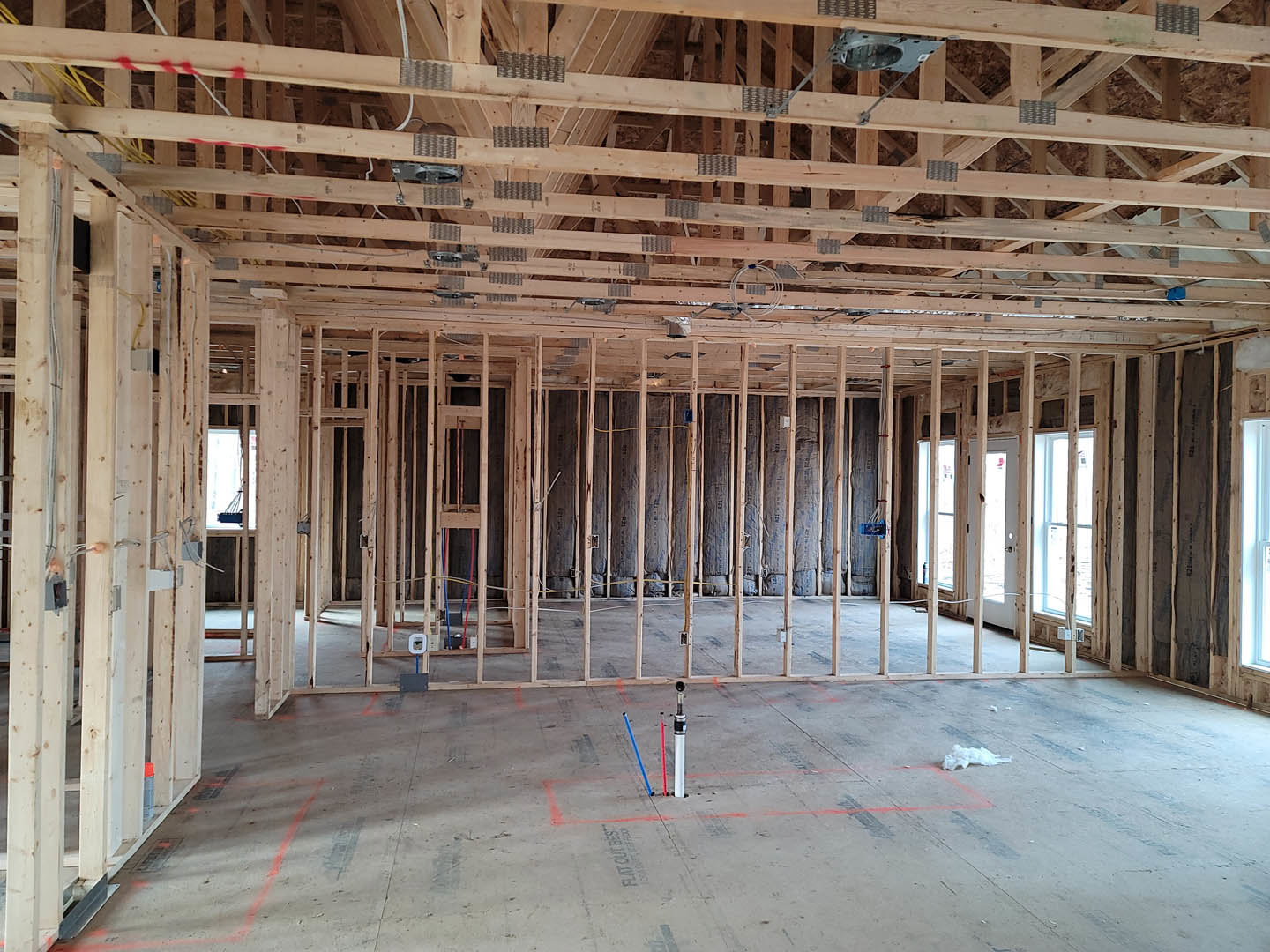Wood-framed house under construction with exposed beams, concrete floor featuring red and blue plumbing pipes, window letting in natural light, white tube with blue end, and blue