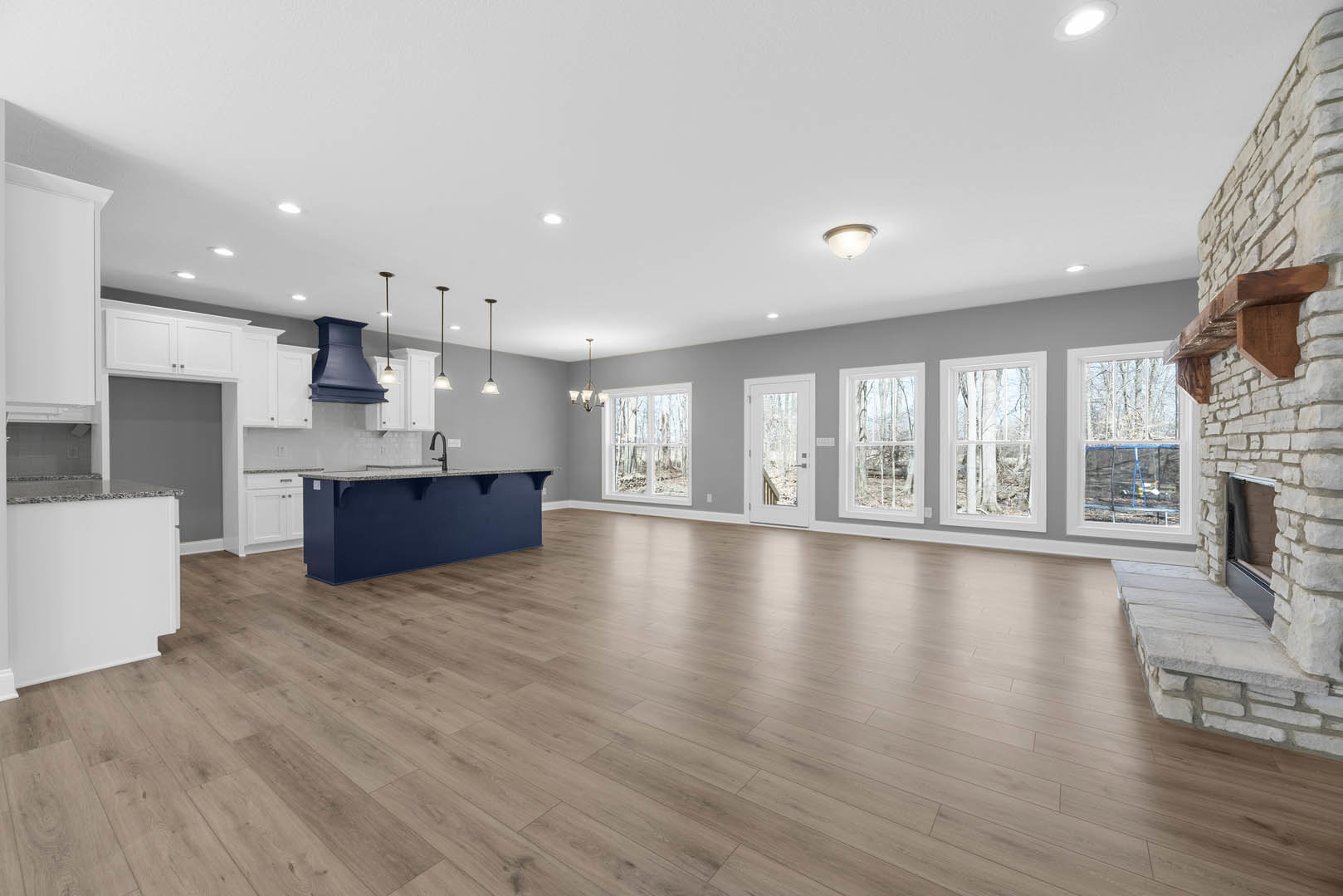 Open-concept kitchen with blue accent wall, central island, hardwood flooring, modern ceiling light fixture, and large windows overlooking forested landscape