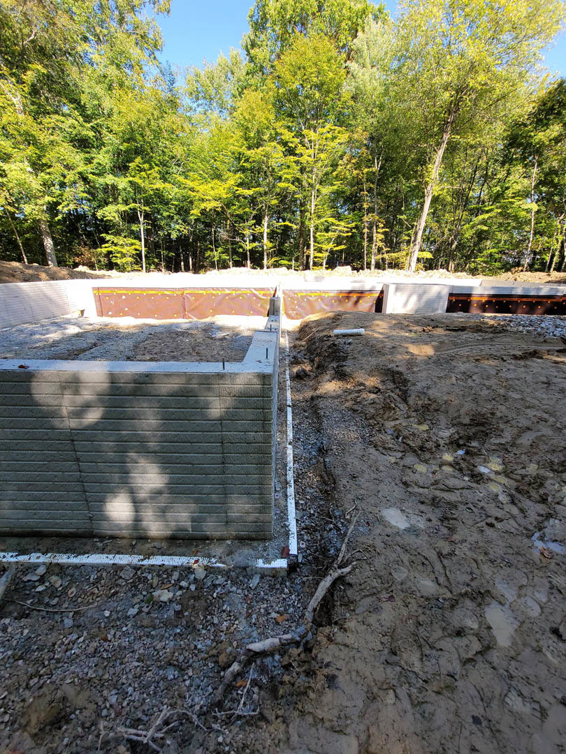 Concrete foundation slab with partial brick wall, muddy ground, and surrounding trees at residential construction site