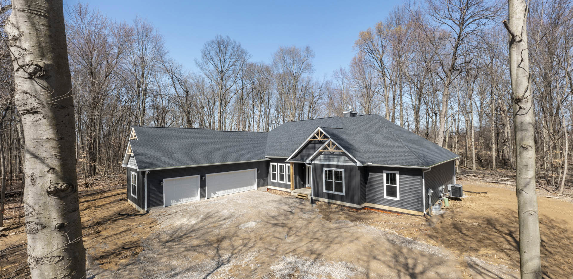 Two-story house with white siding, black roof, attached garage, paved driveway, and mature trees in the background